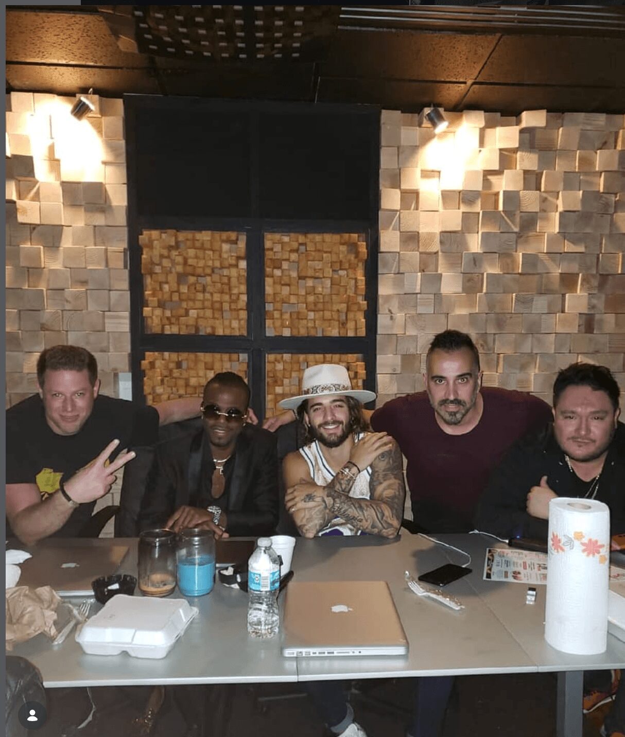 Five people sitting at a table with drinks and items, smiling, in a room with textured wall and lighting.