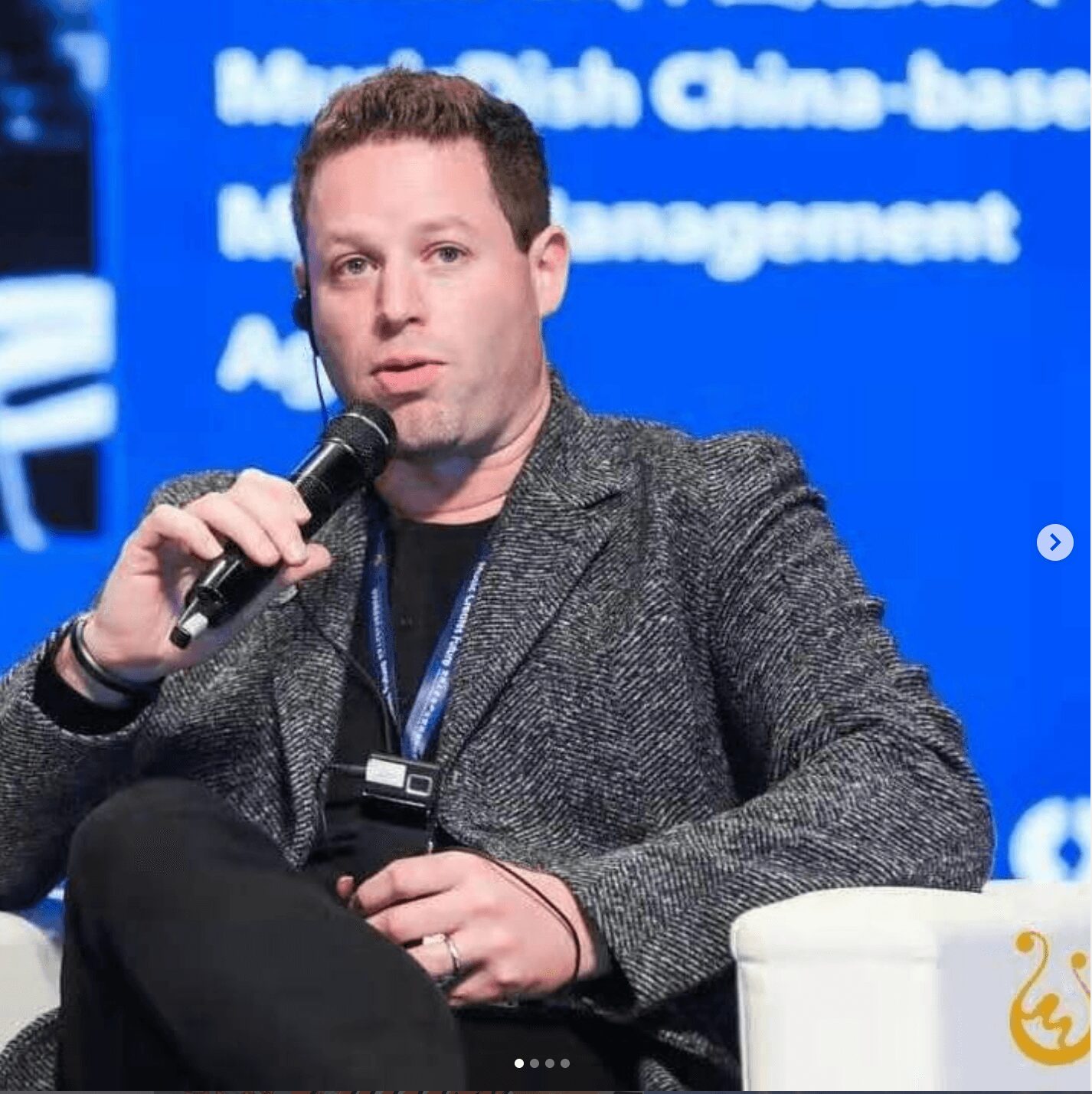 Man holding a microphone and sitting on a white chair at a conference or event.