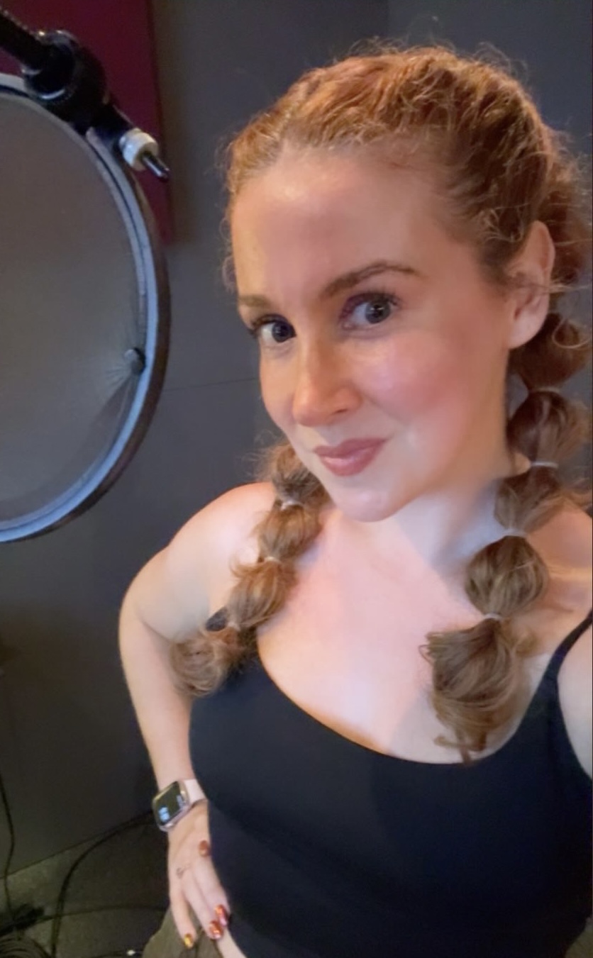 Young woman with braided hair taking a selfie, wearing a black top, in a room with a microphone.