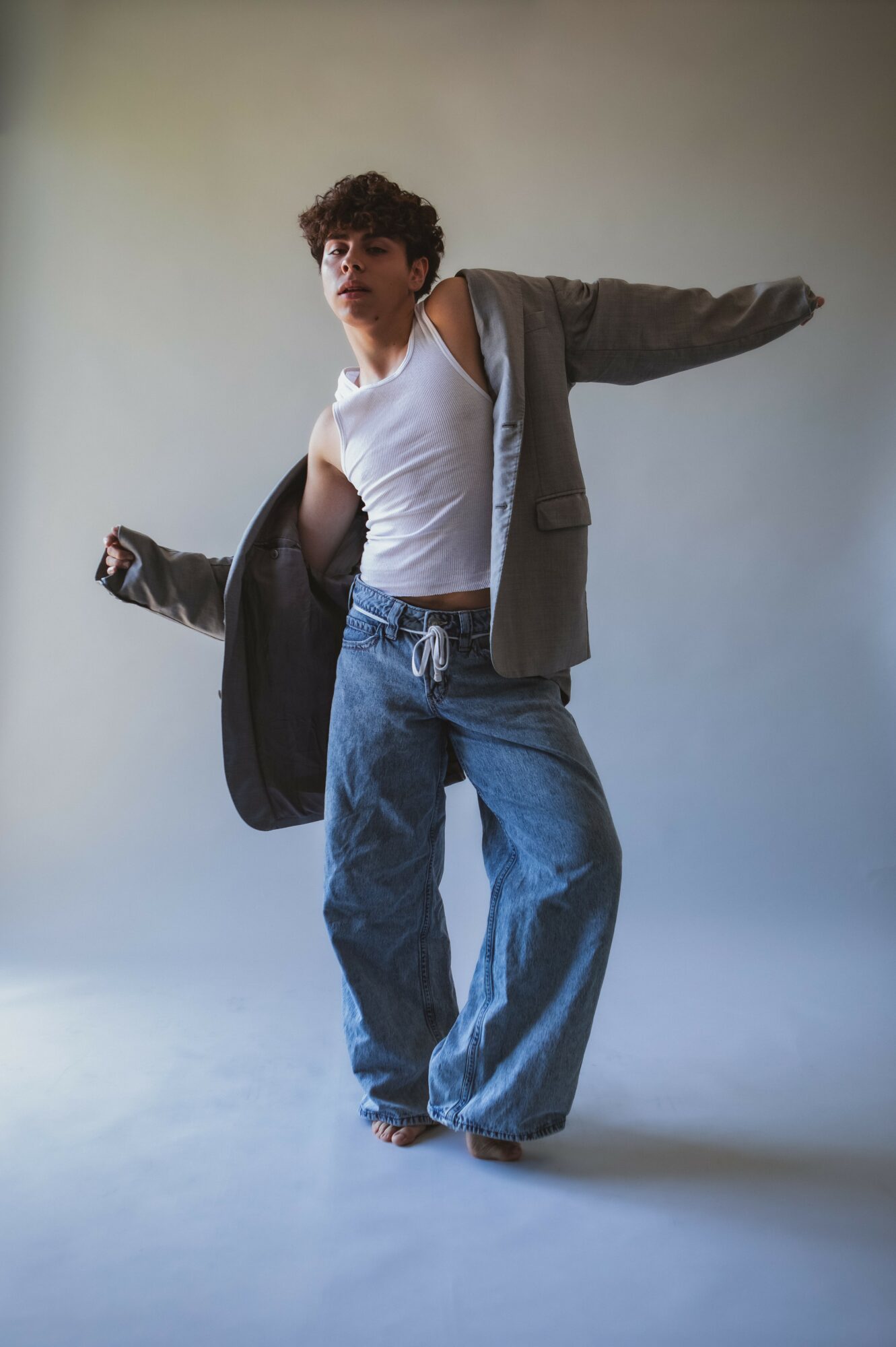 Young person with curly hair in a white tank top, gray jacket, and baggy jeans, posing with arms outstretched.