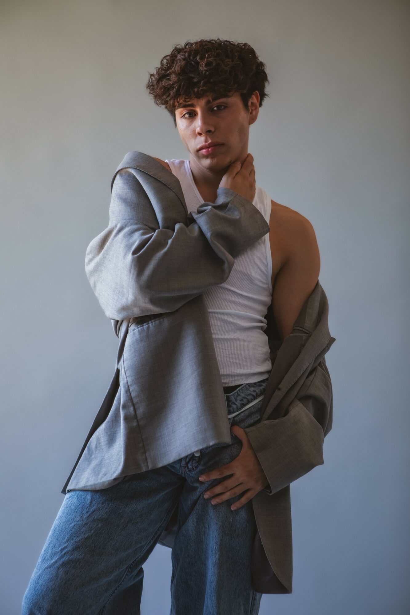 Young person with curly hair wearing a gray jacket, white tank top, and jeans, standing against a plain background.