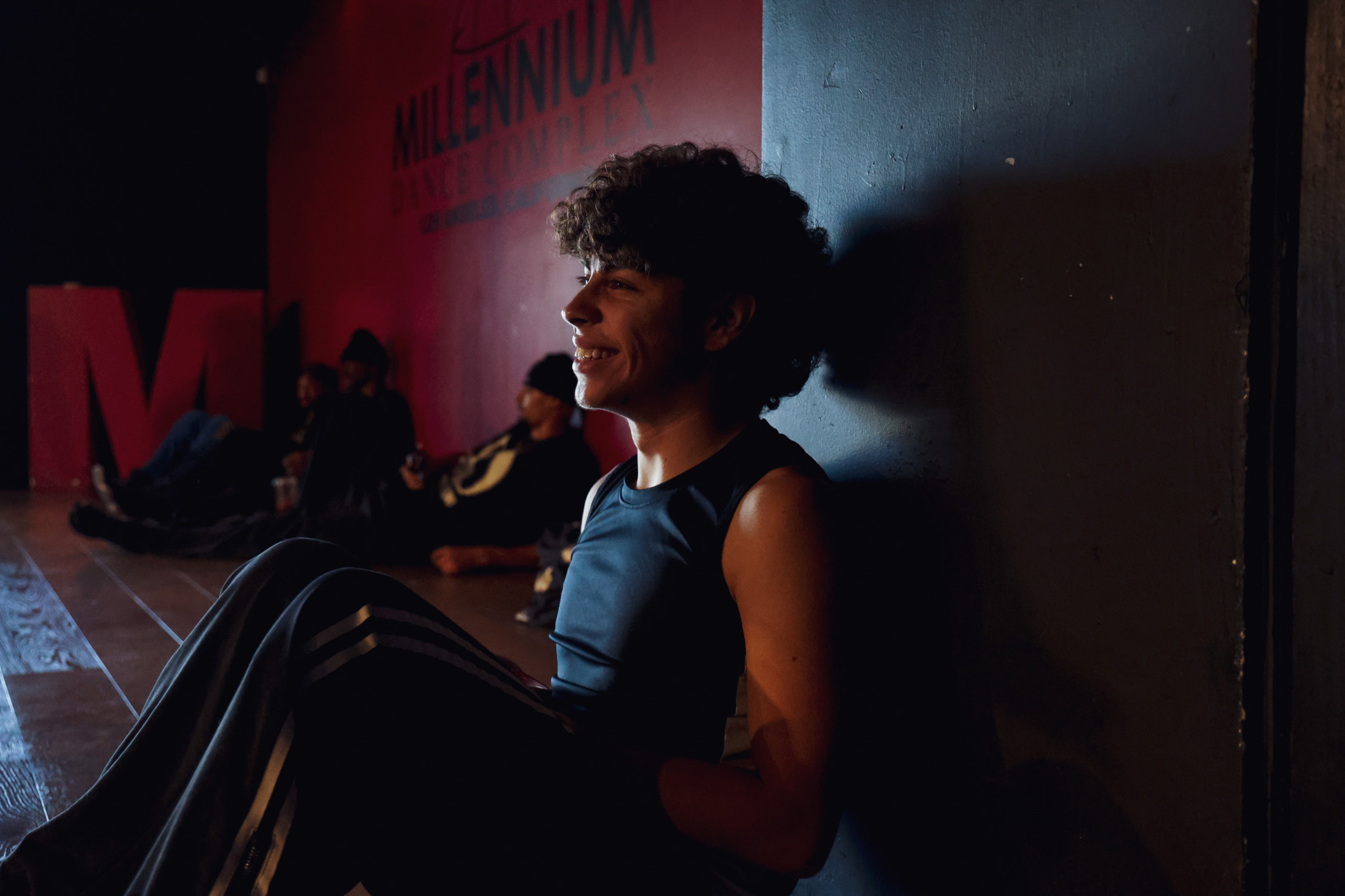 Person with curly hair sitting against a wall, smiling, in a dimly lit room with other people in the background.