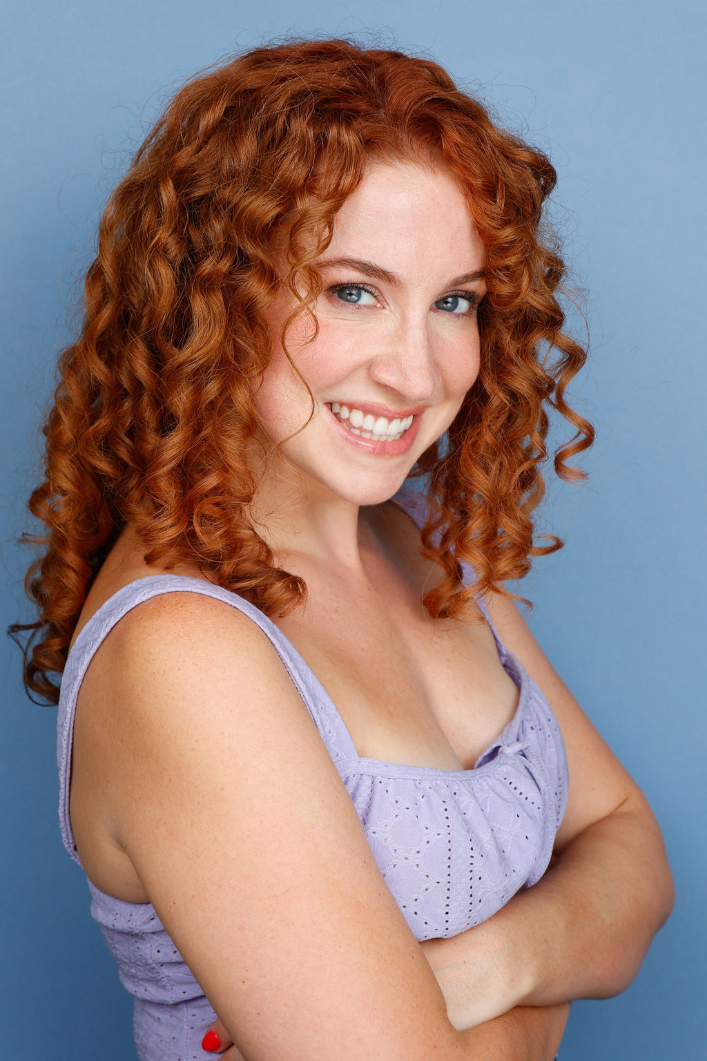Smiling woman with curly red hair wearing a light purple top against a blue background.