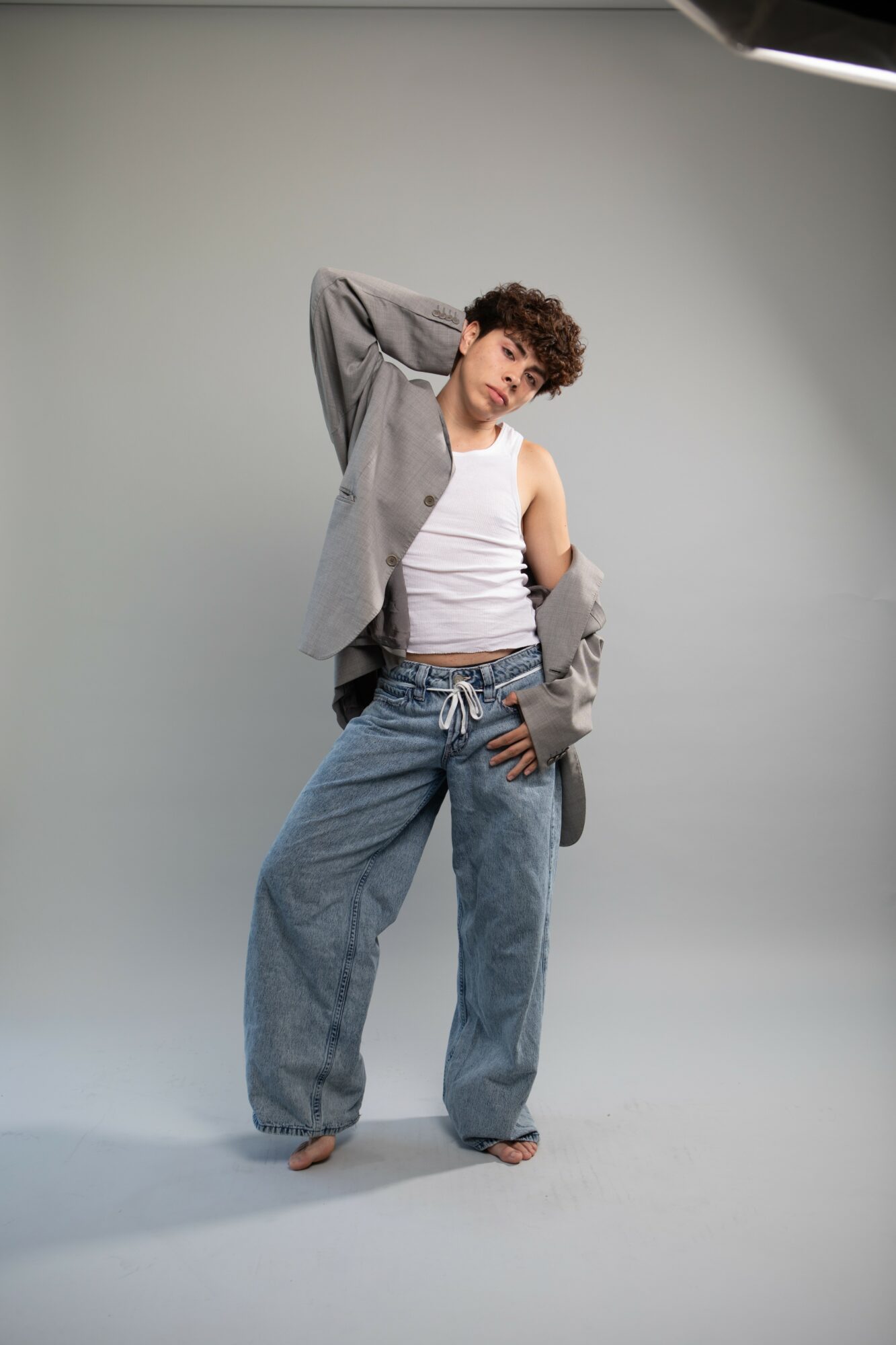 Young person with curly hair poses with one hand behind head, wearing a gray blazer, white tank top, and loose jeans.