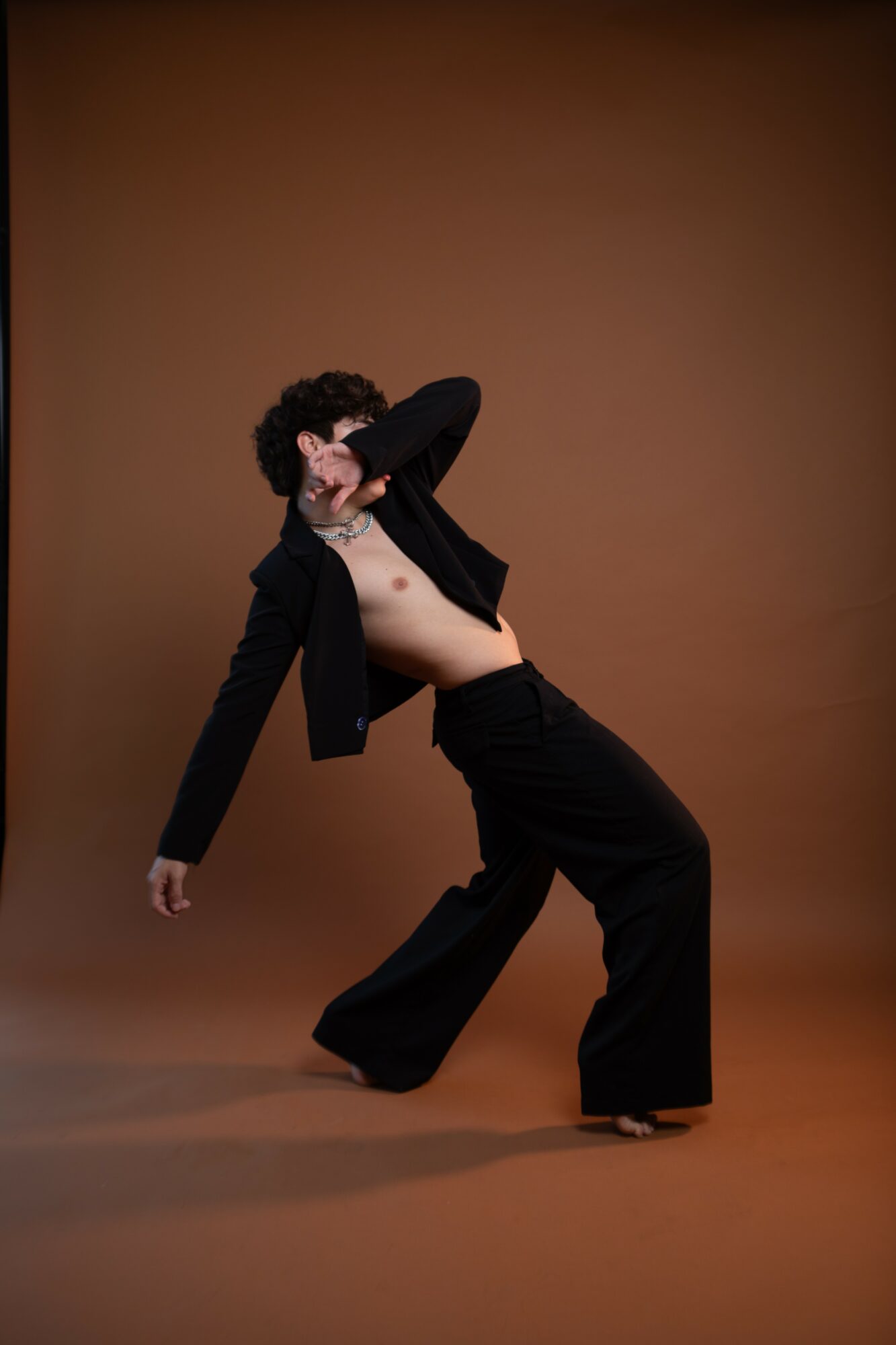 Person with curly hair poses with one arm covering face, wearing black pants and jacket, against brown background.