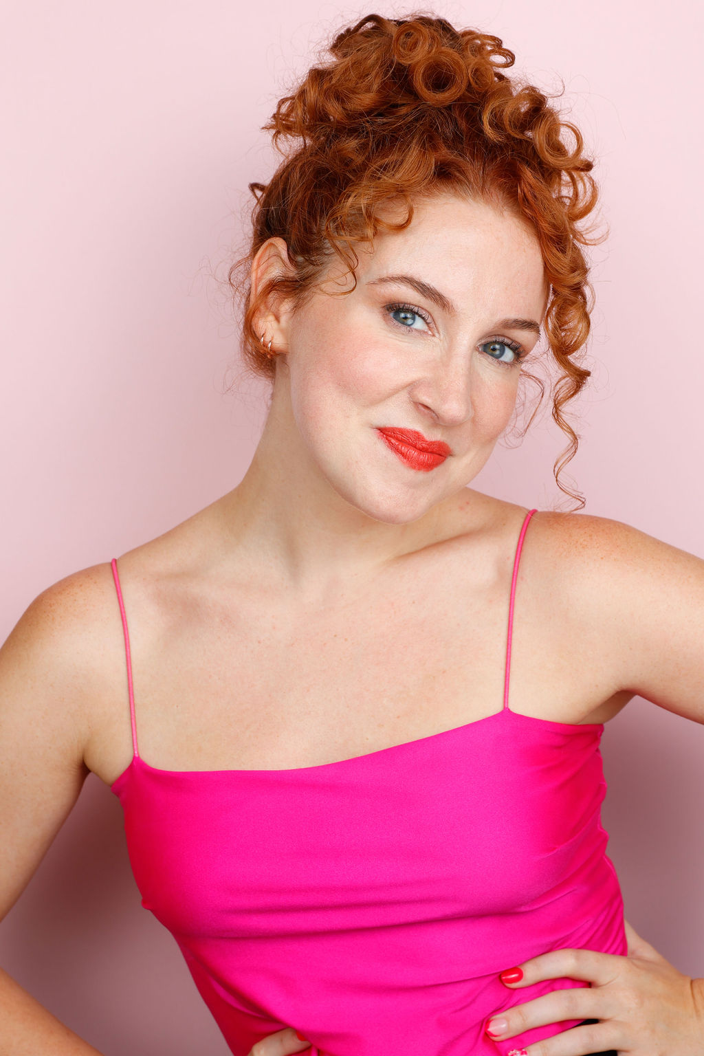 Woman with curly red hair in a pink spaghetti strap top, smiling, against a light pink background.