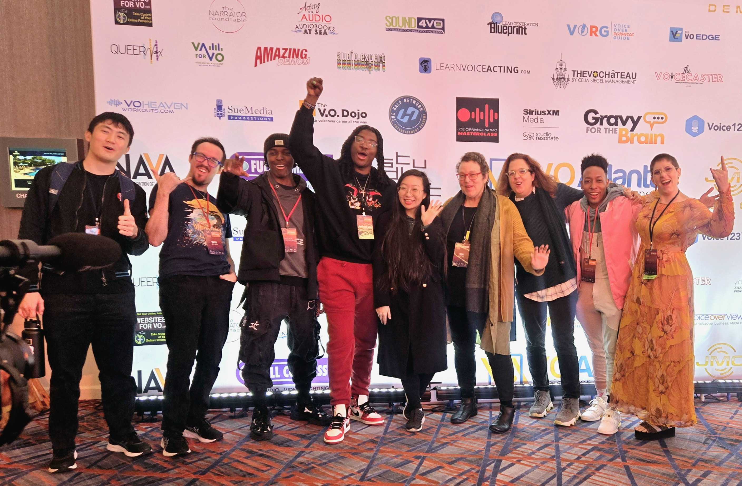 Group of nine people standing on stage in front of a backdrop with logos, some holding microphones and raising hands.