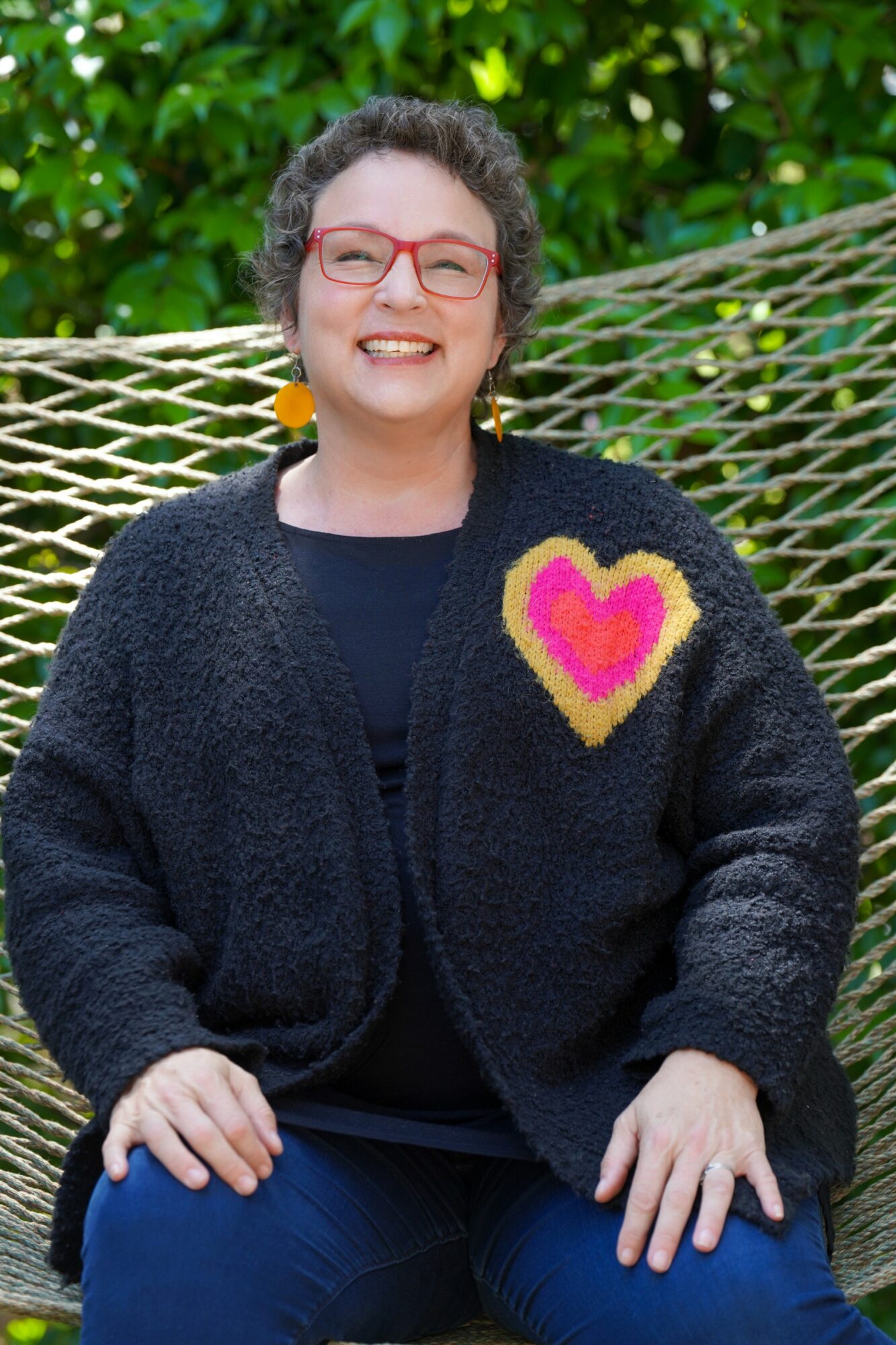 Woman with curly hair, glasses, and earrings smiling outdoors, wearing a black cardigan with a colorful heart patch.