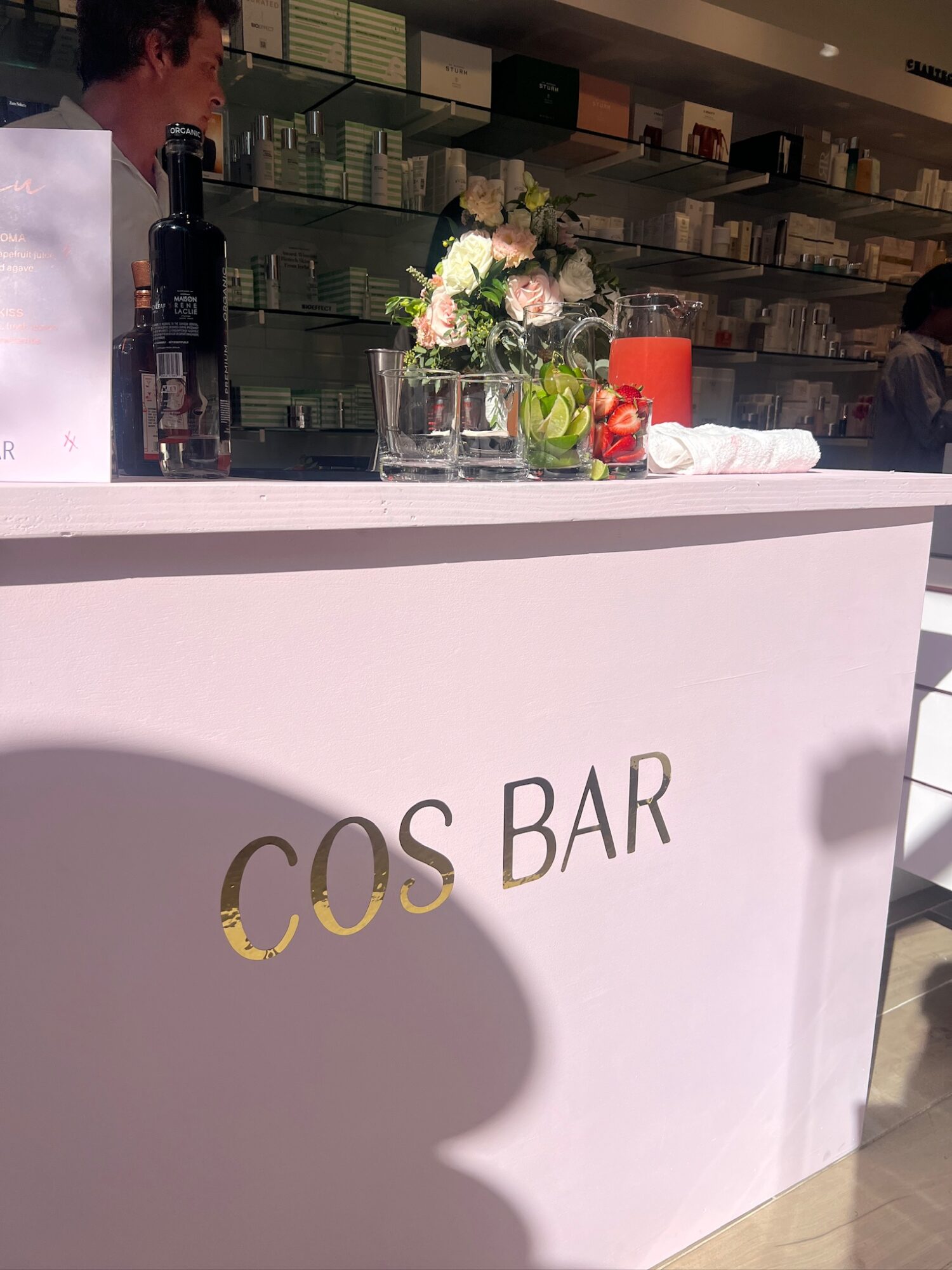 Bar counter with flowers, a red container, and a white cloth, with shelves in the background.