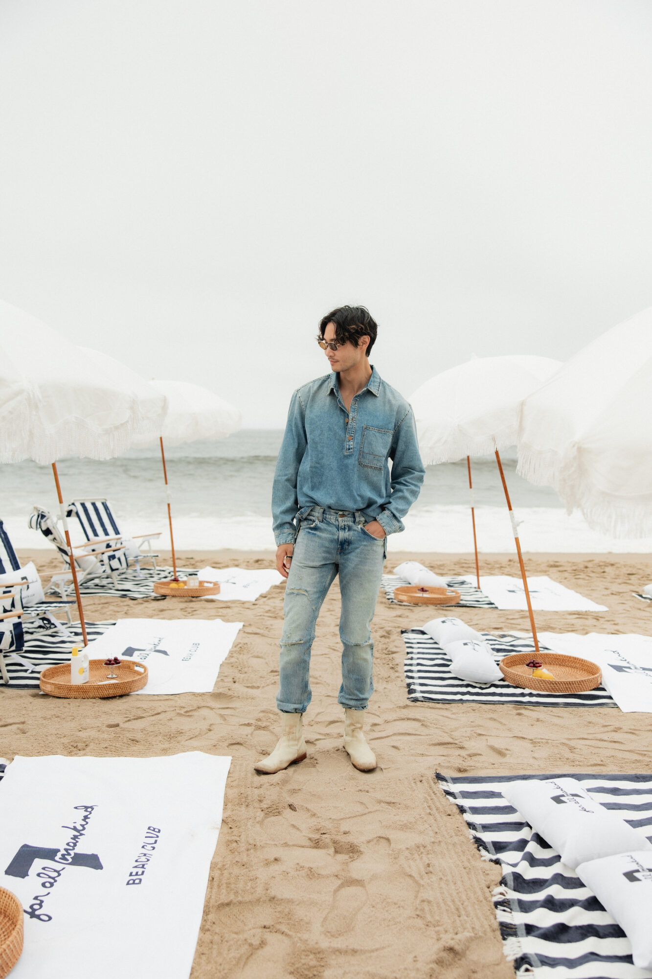Young man in denim outfit standing on sandy beach with striped towels and umbrellas nearby.