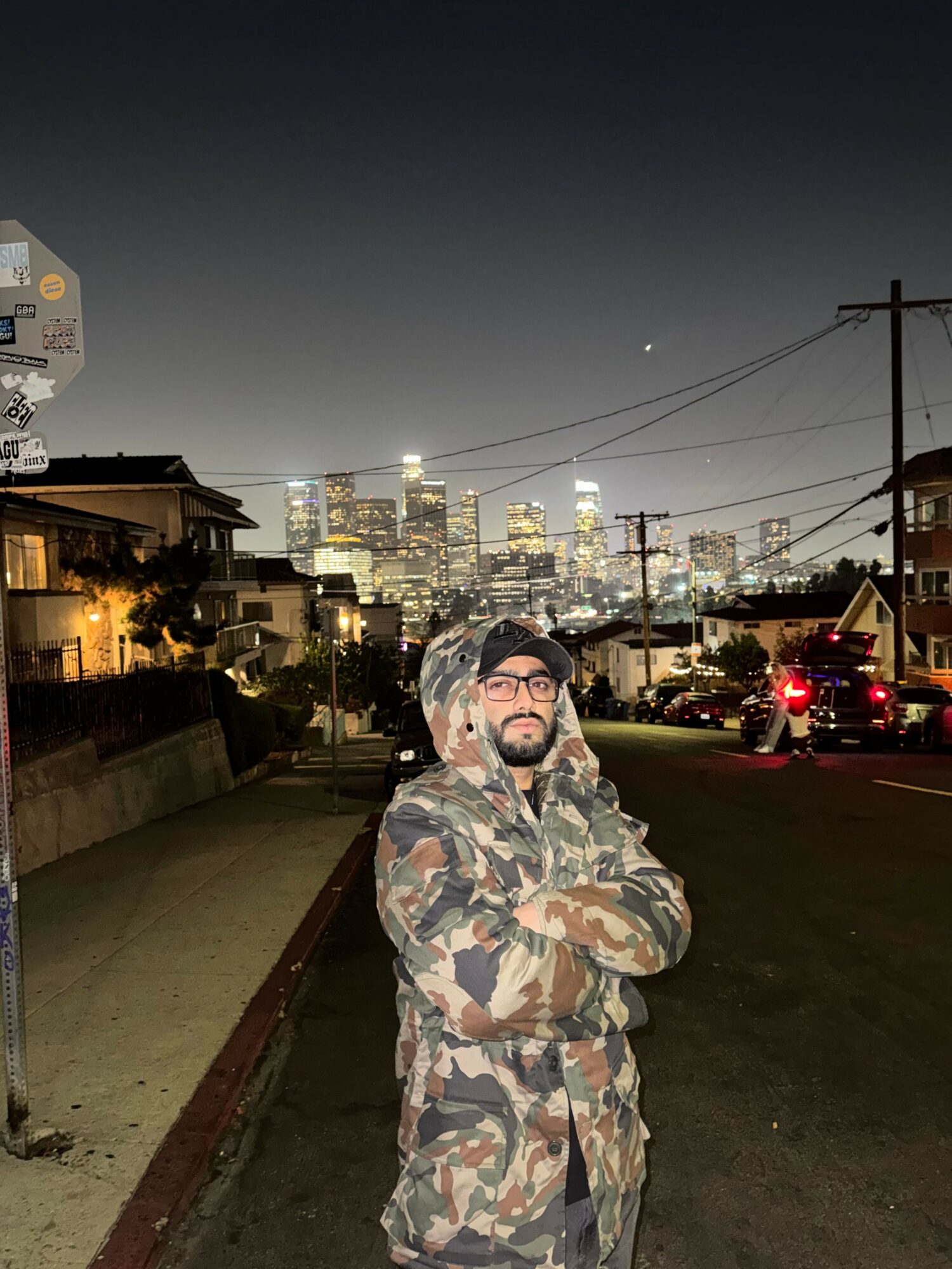 Man in camouflage jacket and glasses standing on sidewalk at night with city skyline and streetlights behind him.