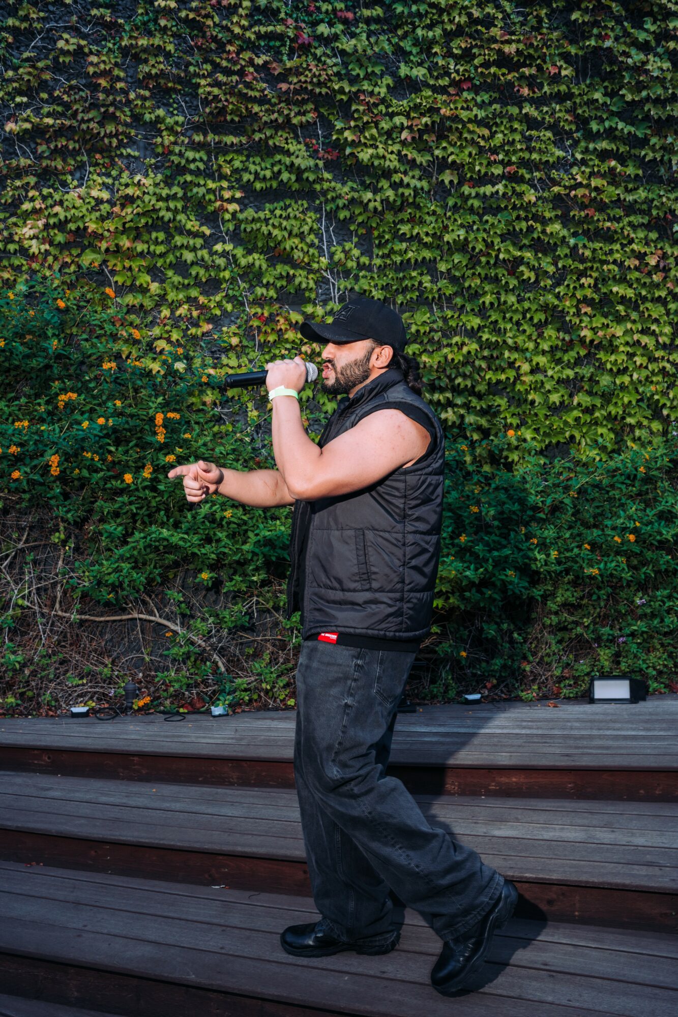 Person standing outdoors in front of a dense green hedge, wearing a black vest, pants, and cap, looking at a watch.