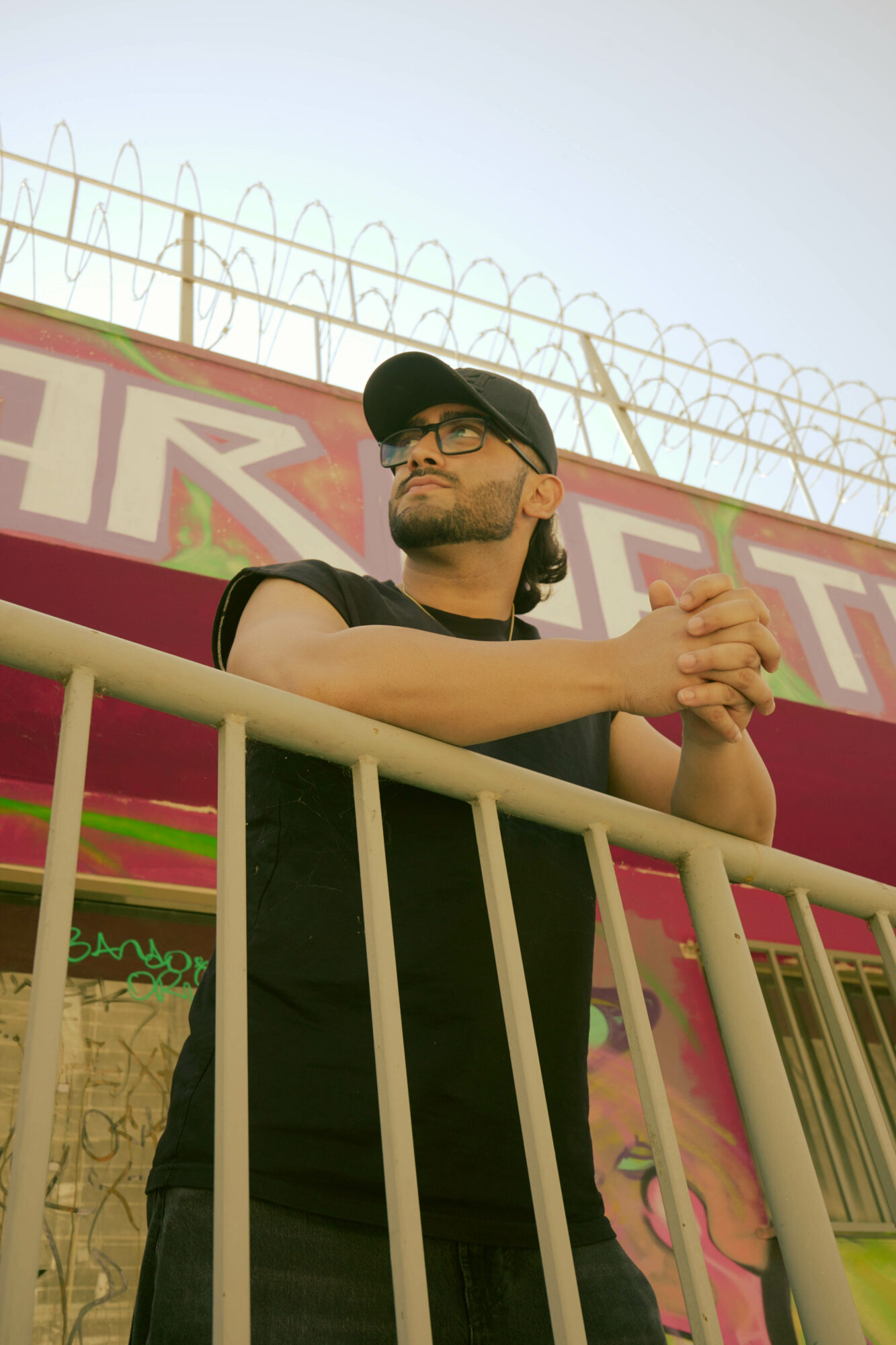 Man wearing glasses and a cap leaning on a railing, with a colorful background and barbed wire above.