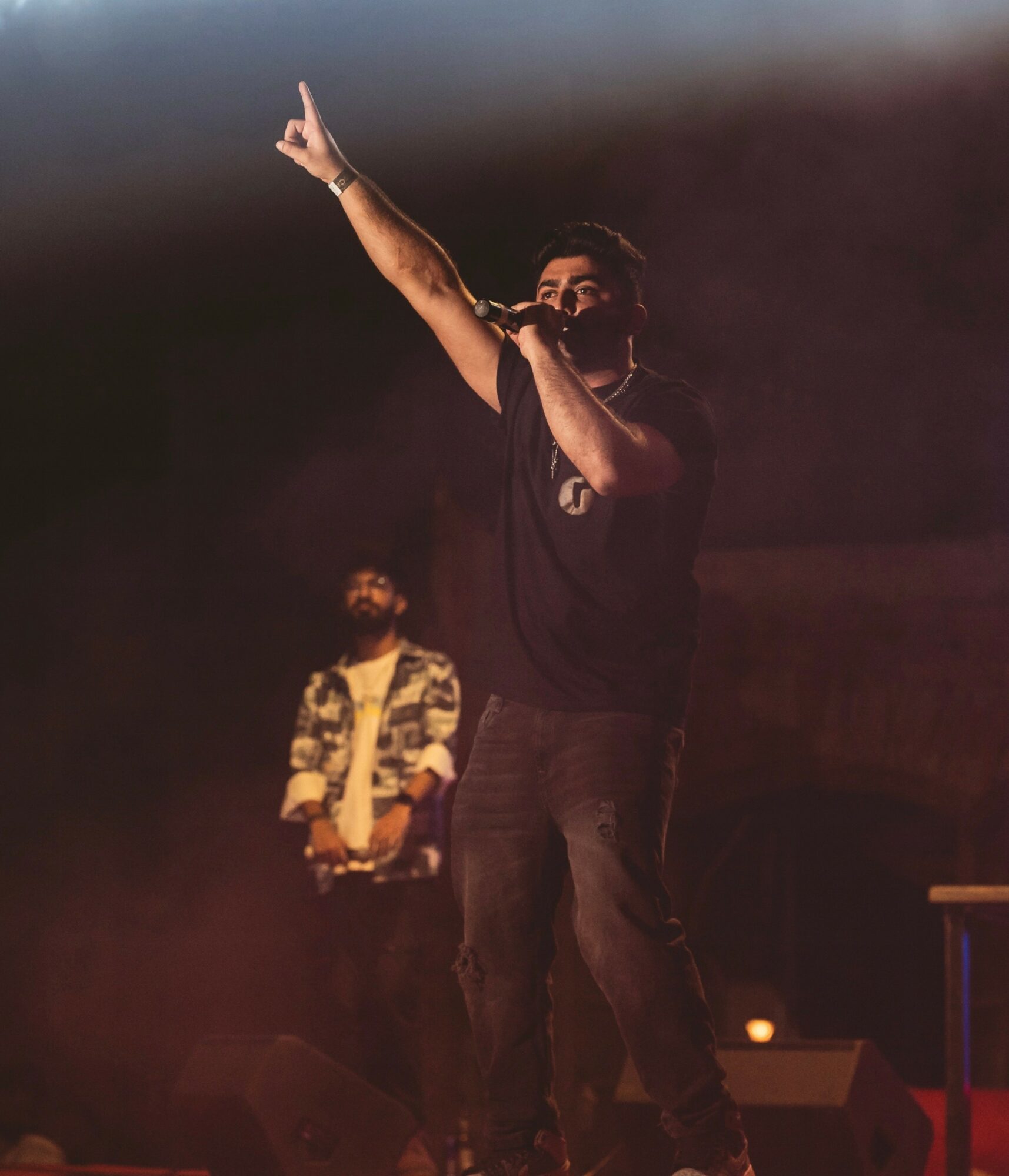 Performer on stage with one arm raised and holding a microphone, another person in background, dark stage lighting.