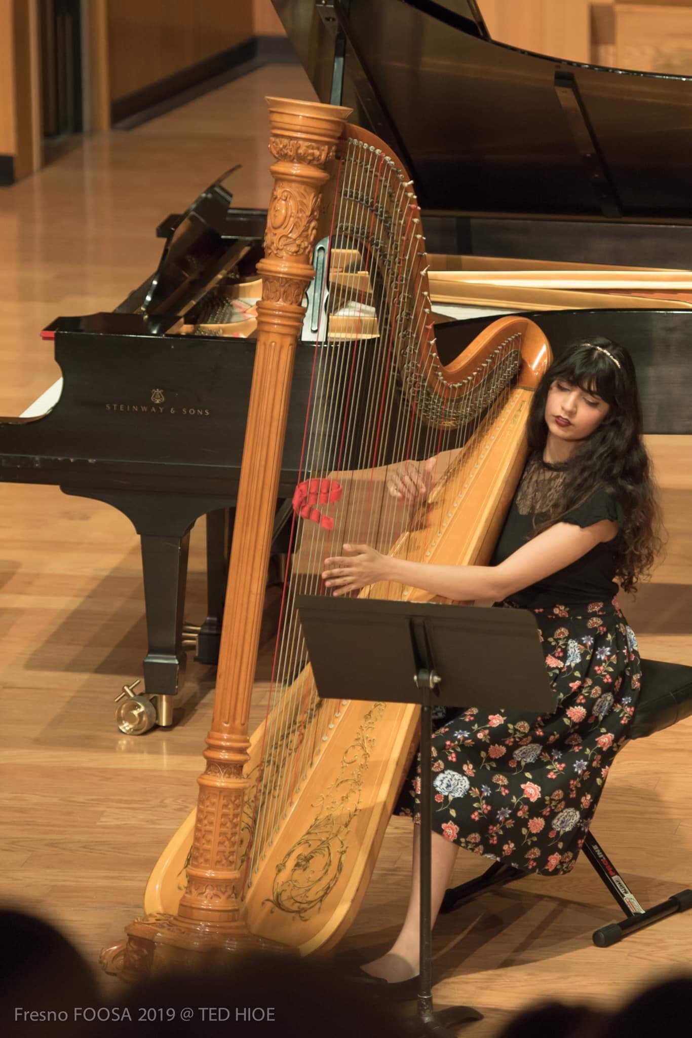 Young woman with long dark hair playing a harp on stage, with a grand piano in the background.