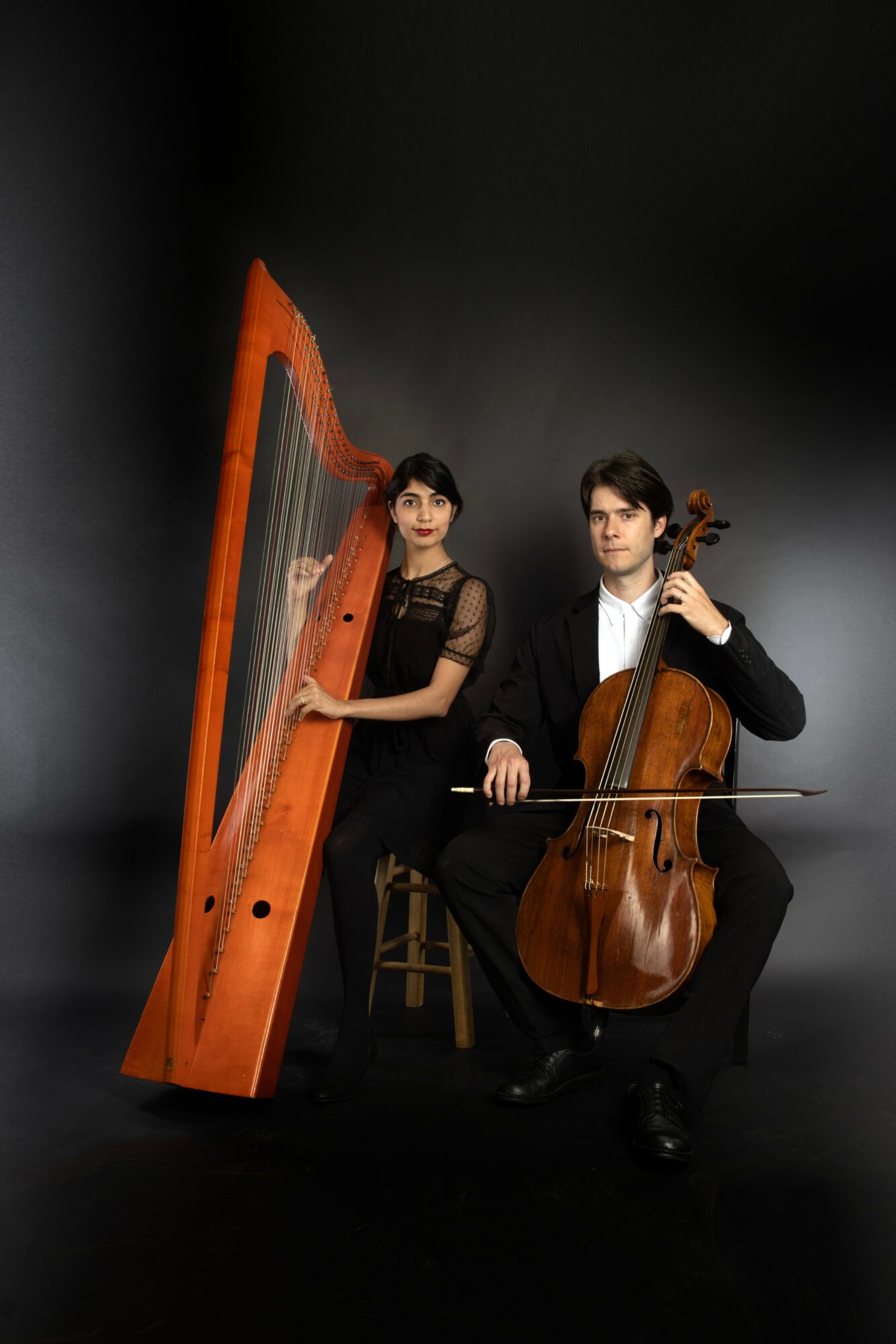 Two musicians, a woman and a man, with a harp and a cello, seated against a dark background.