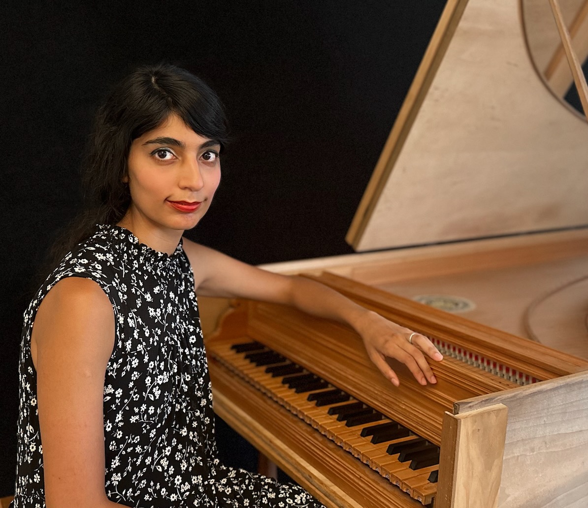 Woman with dark hair in a floral dress sitting at a wooden piano, touching the keys.