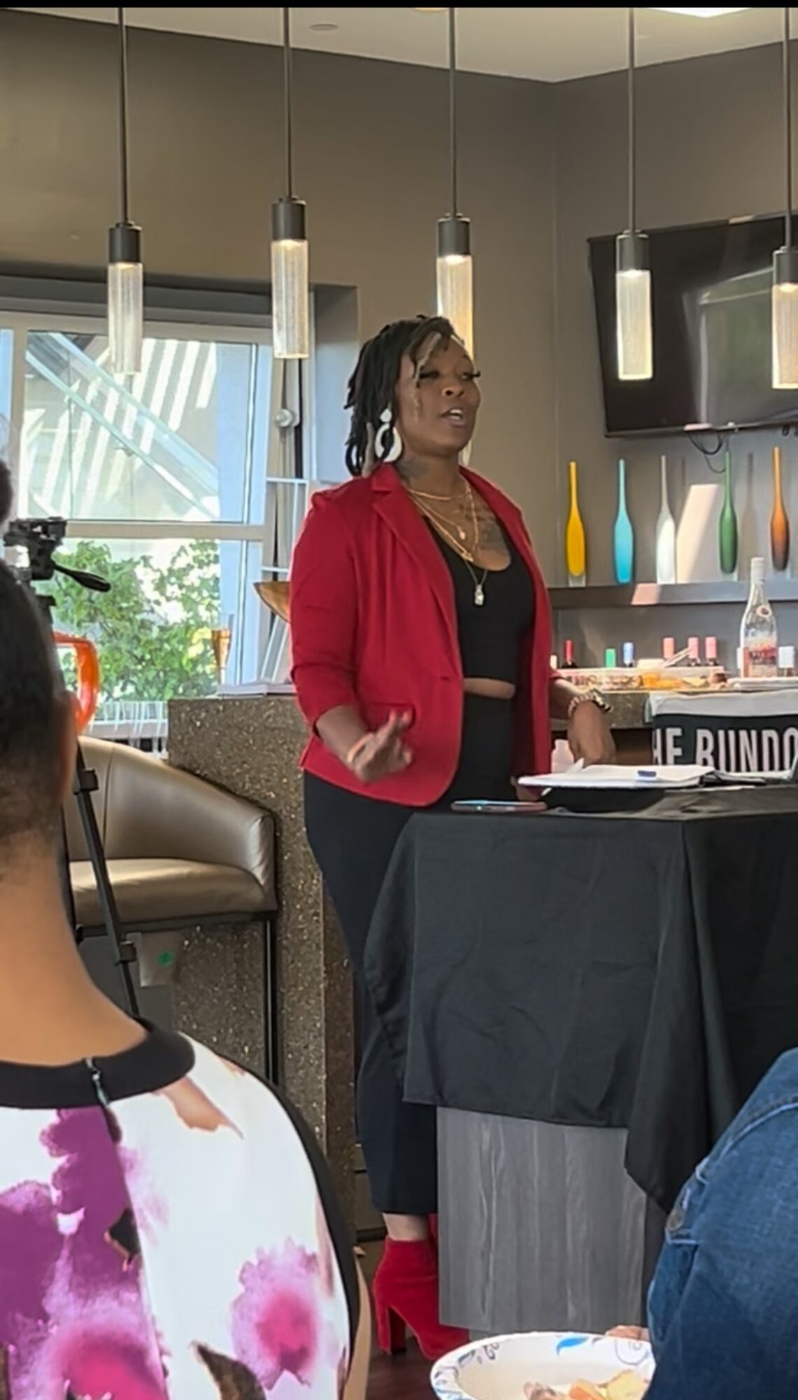 Woman in red jacket speaking in a modern kitchen with hanging lights and colorful utensils.