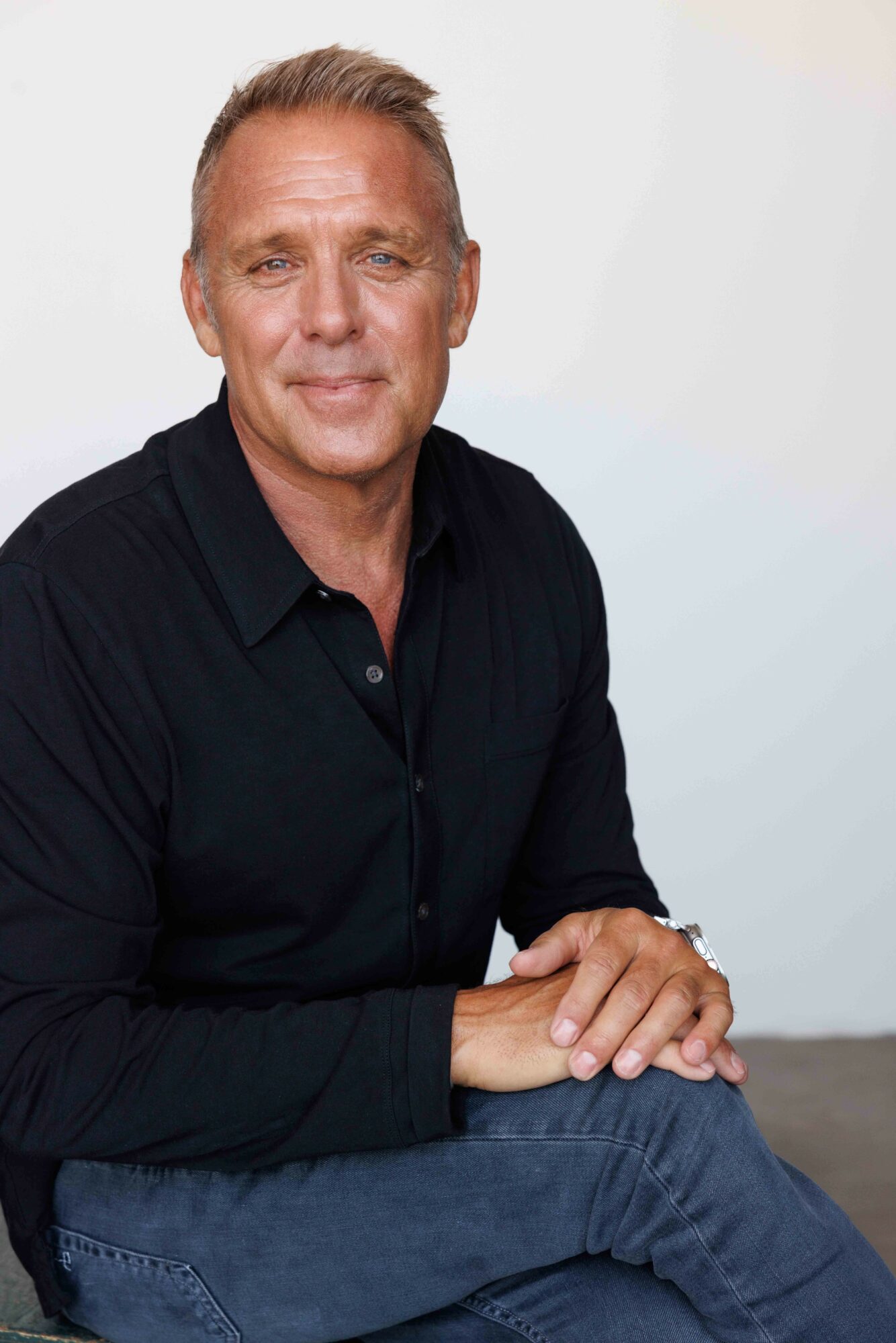 Middle-aged man with short hair, wearing a black shirt, sitting with hands folded, smiling against a plain background.
