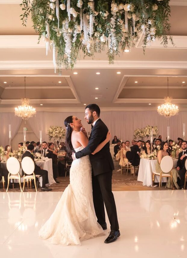 Couple dancing in a decorated ballroom with chandeliers and seated guests in the background.