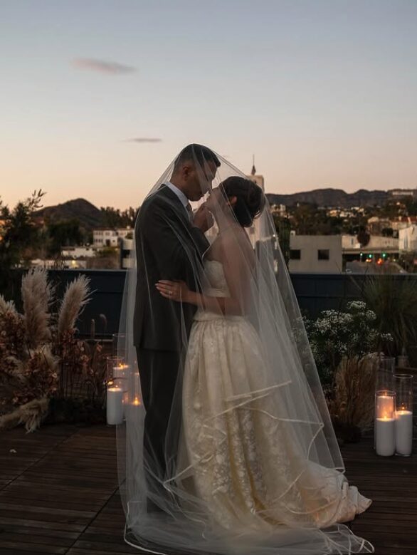 Bride and groom standing close together on a rooftop at sunset, with candles and plants around them.