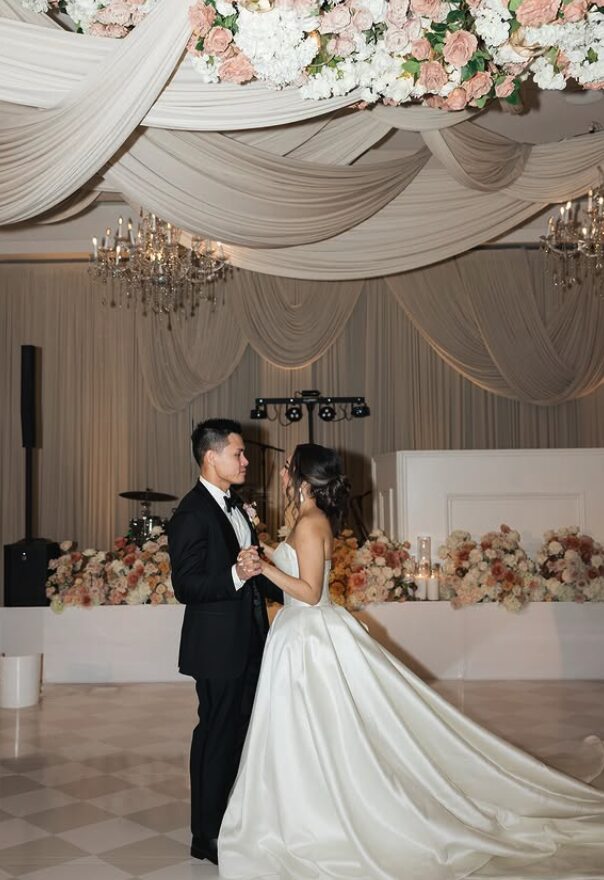 Bride and groom holding hands and looking at each other in a decorated wedding hall with floral arrangements and chandeliers.
