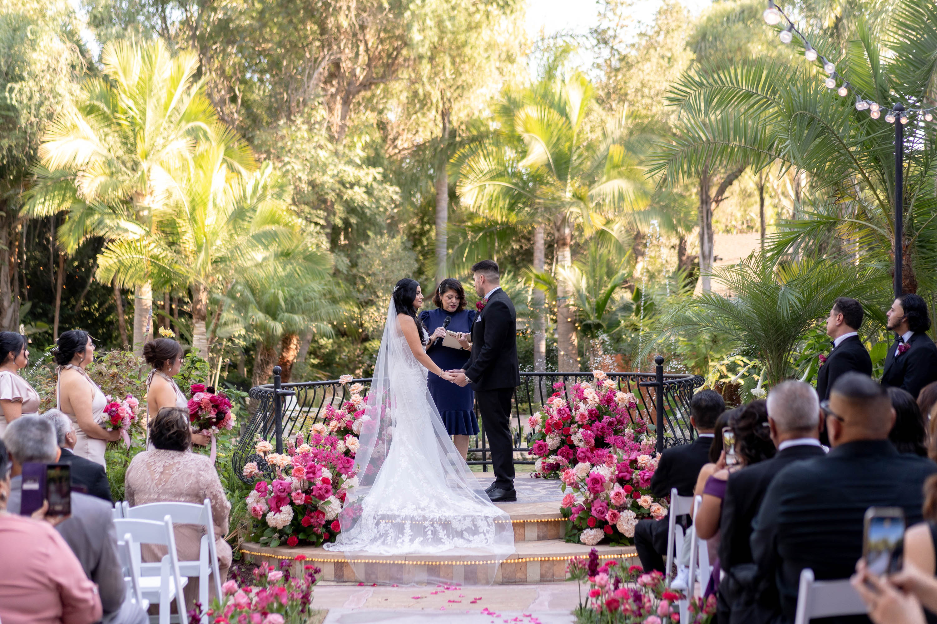 Bride and groom exchange vows outdoors surrounded by guests and pink floral arrangements, with lush greenery and palm trees in background.