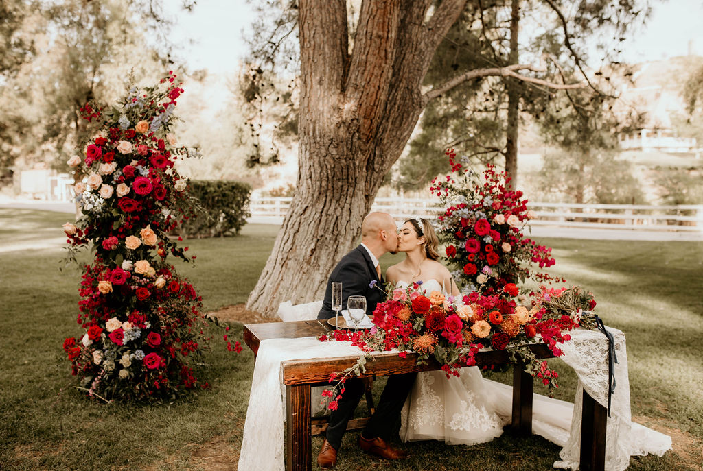 Bride and groom sitting at a table outdoors with large floral arrangements and a tree in the background.