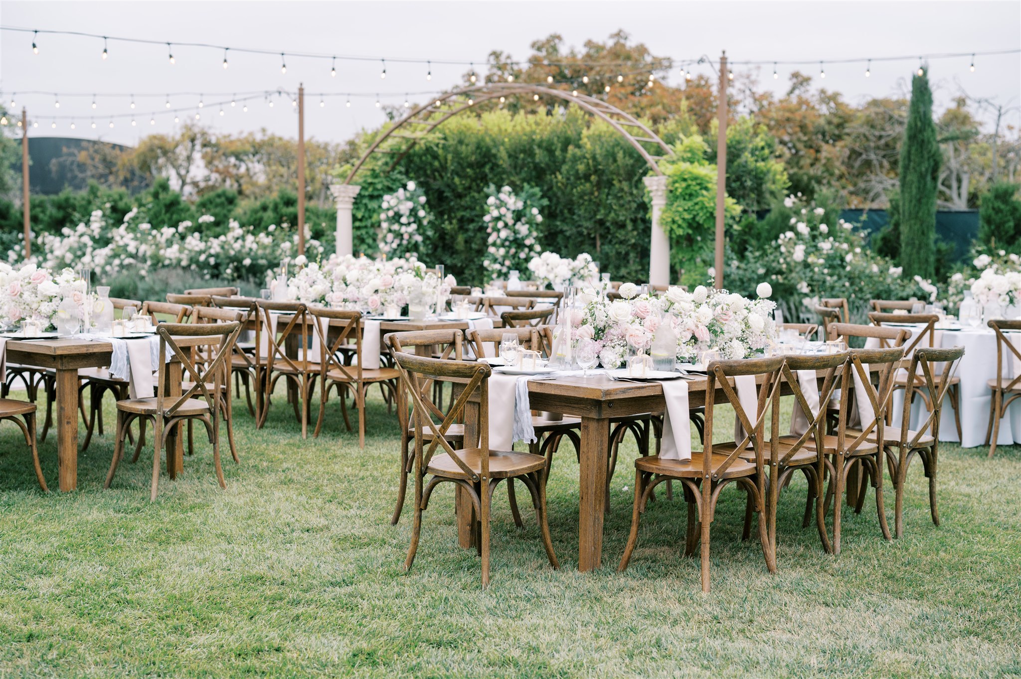 Outdoor event setup with wooden tables, white tableware, and chairs on a grassy area with floral decorations and greenery.