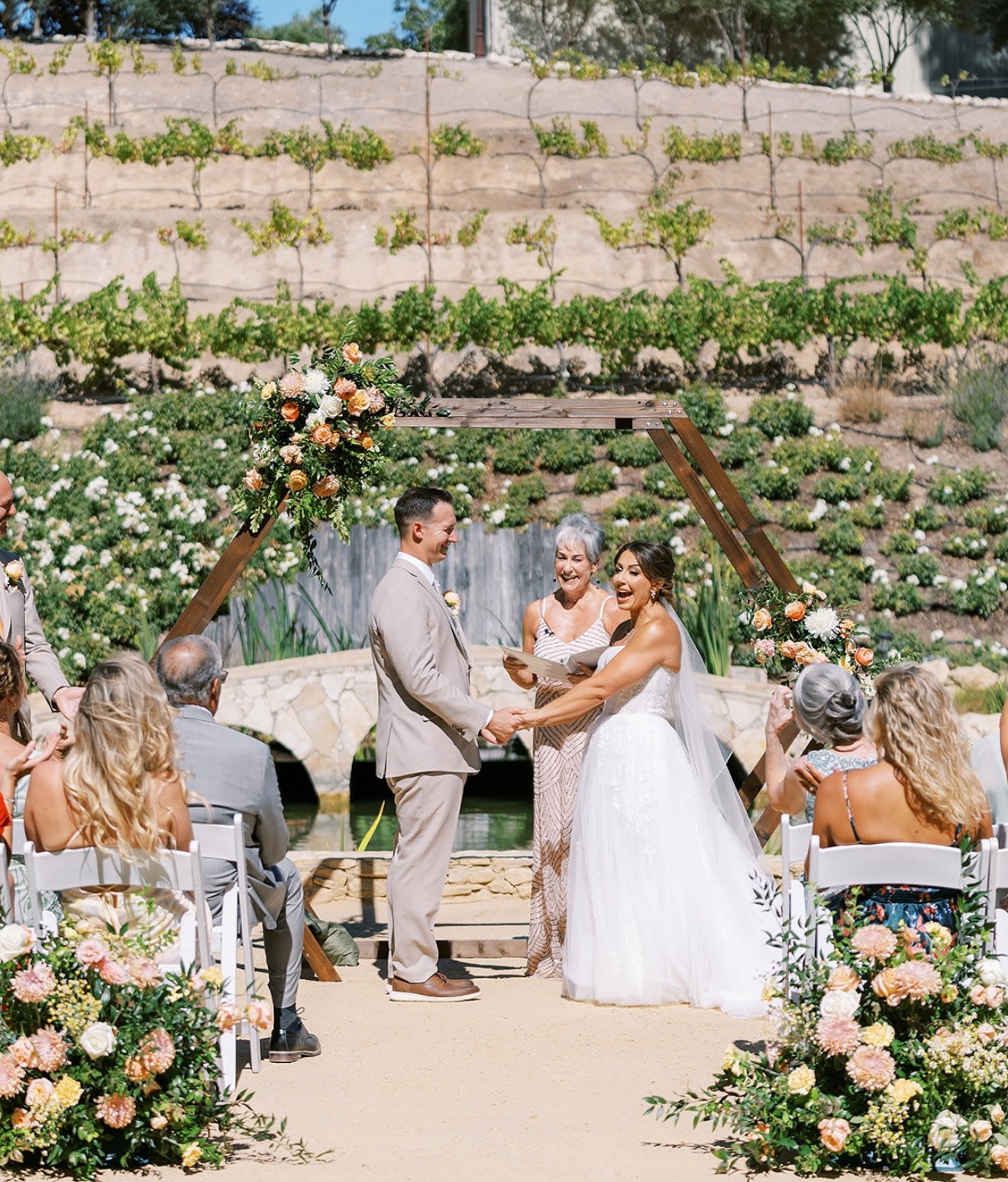 Wedding ceremony with couple, officiant, and guests outdoors, surrounded by flowers and greenery.