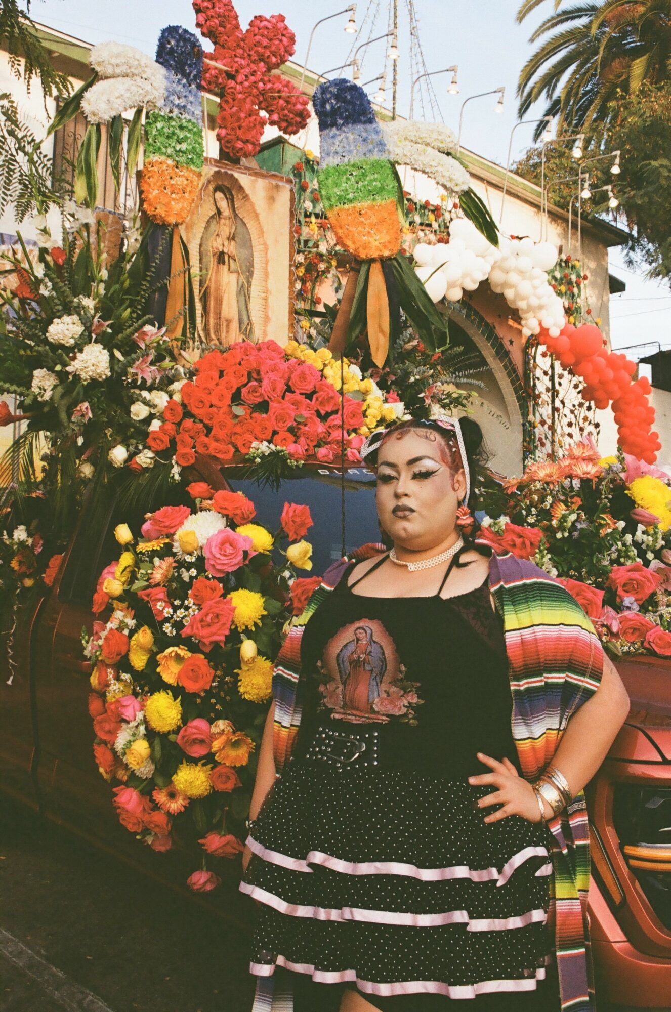 Woman in black dress with floral patterns, standing among colorful flowers and decorations, holding her waist with one hand.