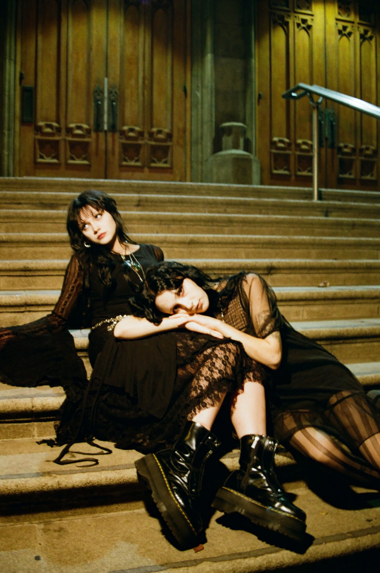 Two women in black outfits and boots sit on wooden stairs inside a building with wooden paneling and a railing.