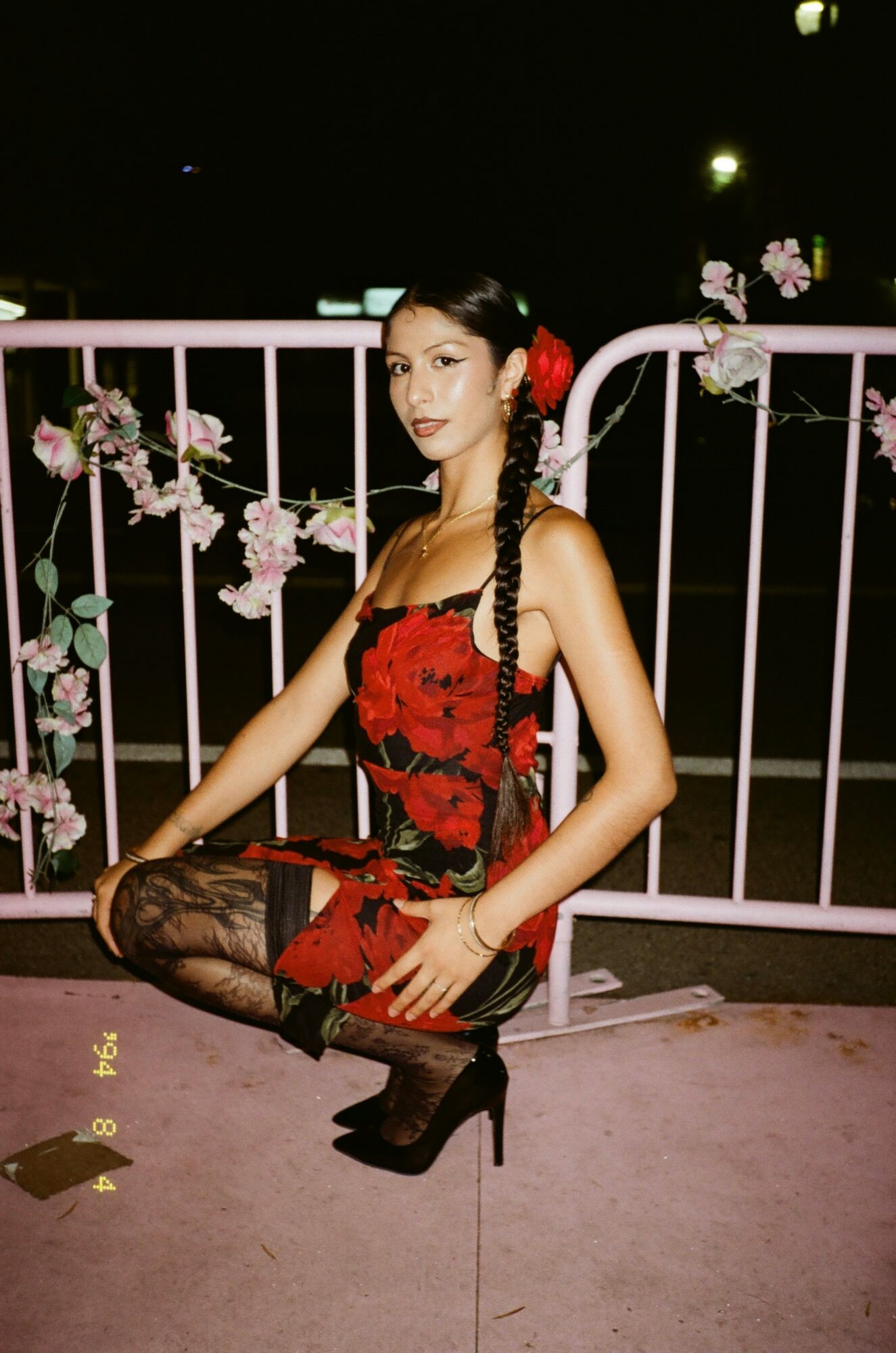 Young woman in red and black dress with floral pattern, black lace stockings, high heels, and braided hair with red flower, squatting outdoors at night.