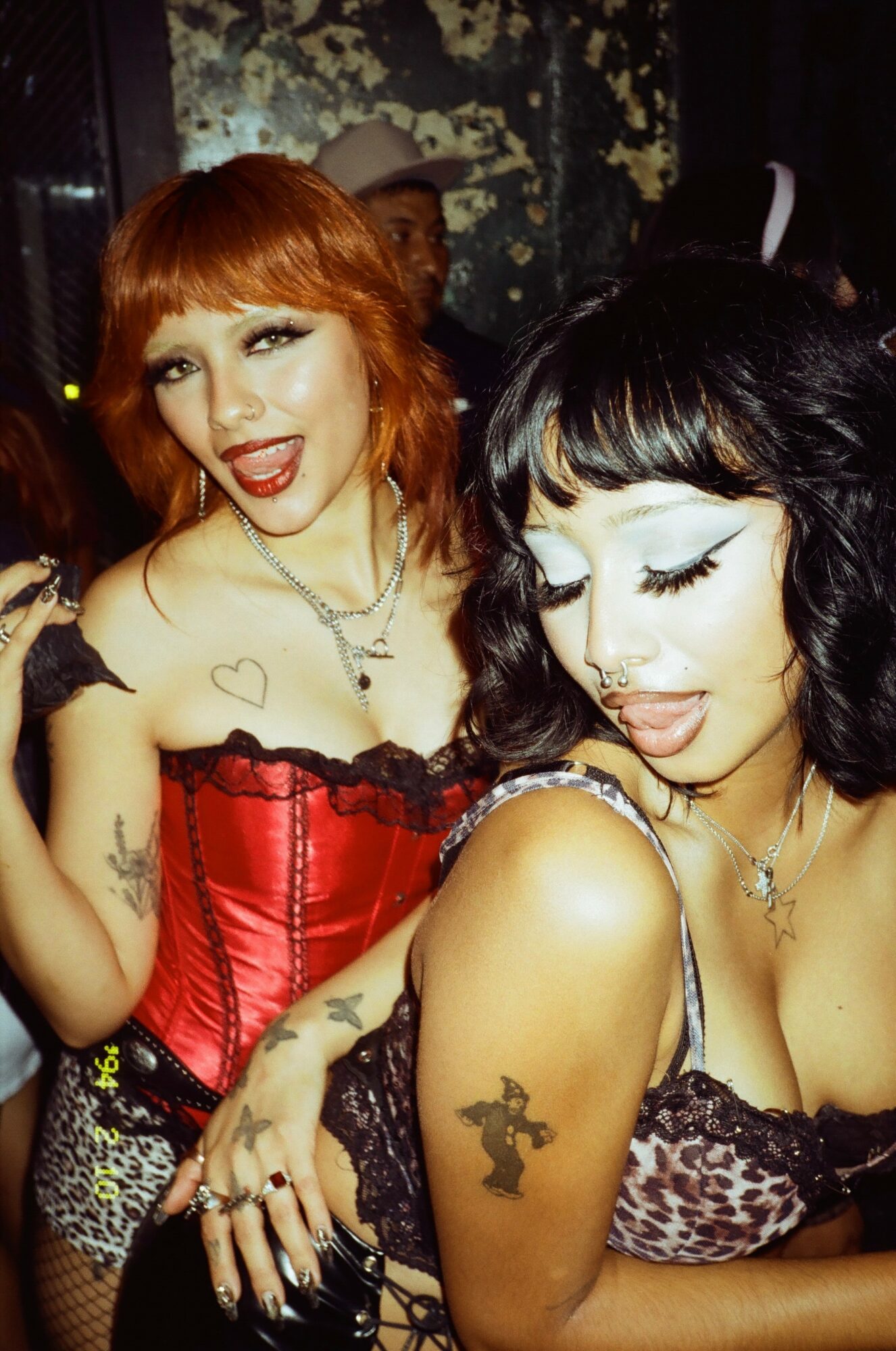 Two women with tattoos and styled hair pose together, one with red hair and the other with black hair, in a dimly lit setting.
