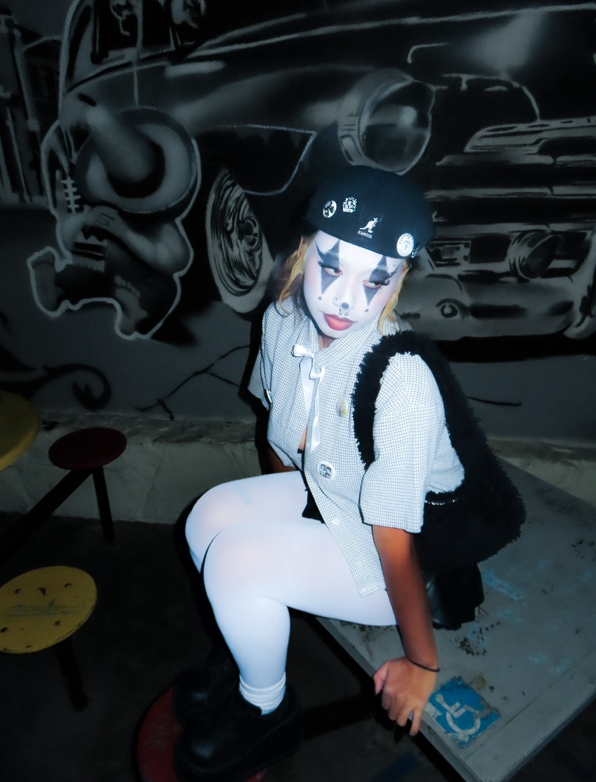Person dressed as a mime with face paint, sitting on a stool, in front of a car mural background.