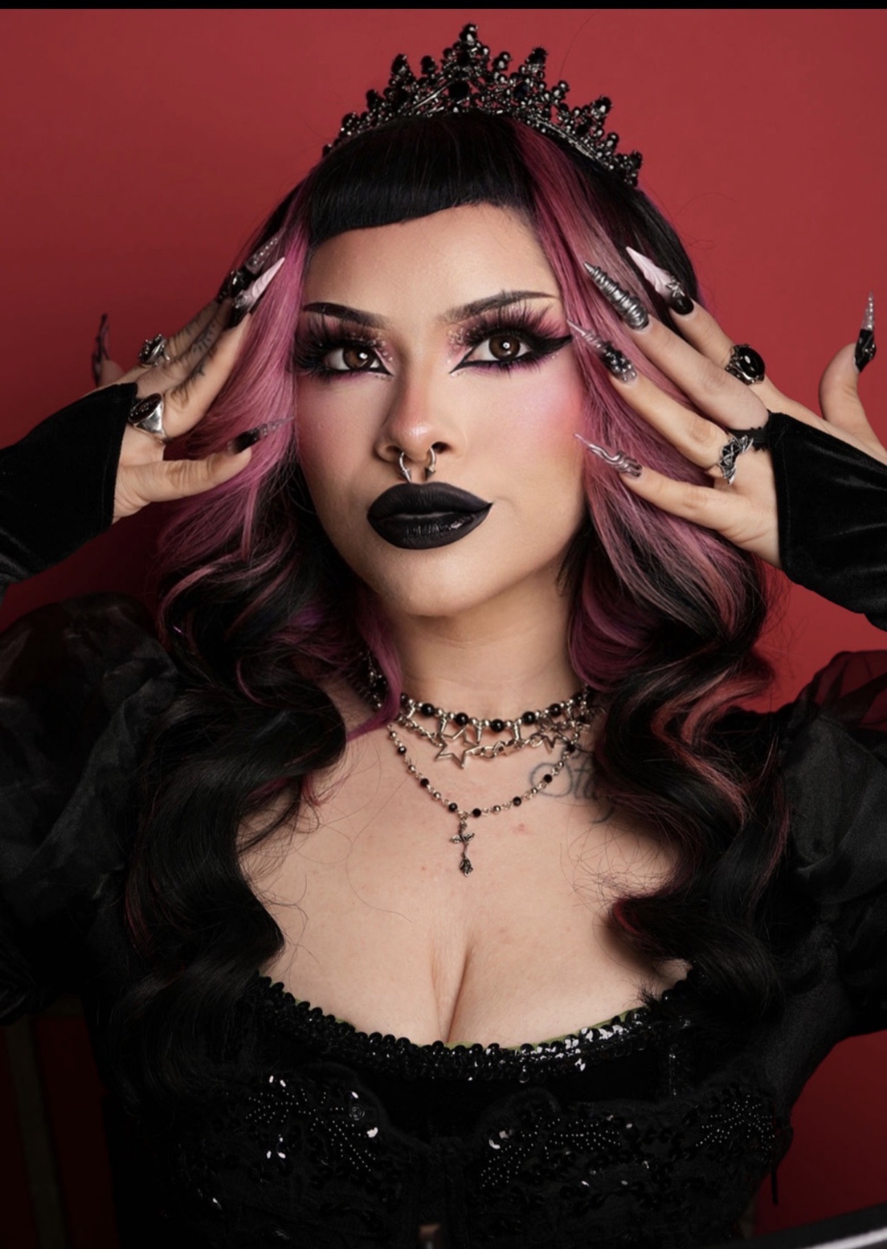Woman with dark makeup, black crown, and jewelry poses against red background.