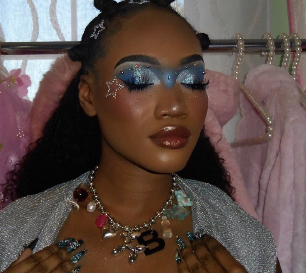 Person with blue glittery eye makeup, star-shaped face decoration, and jewelry, in front of pink clothing and accessories.
