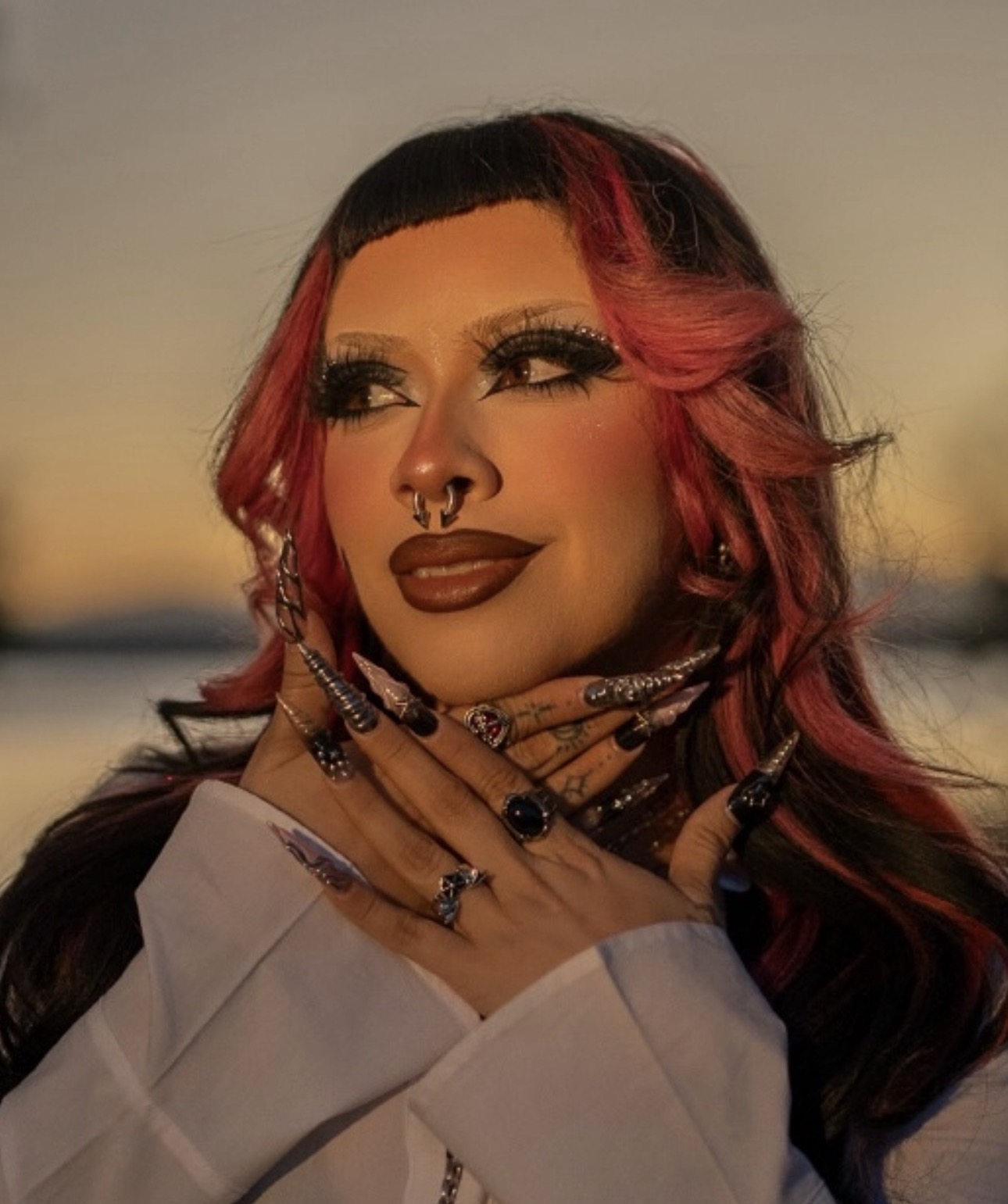 Woman with pink and black hair, makeup, and multiple rings, posing outdoors at sunset.
