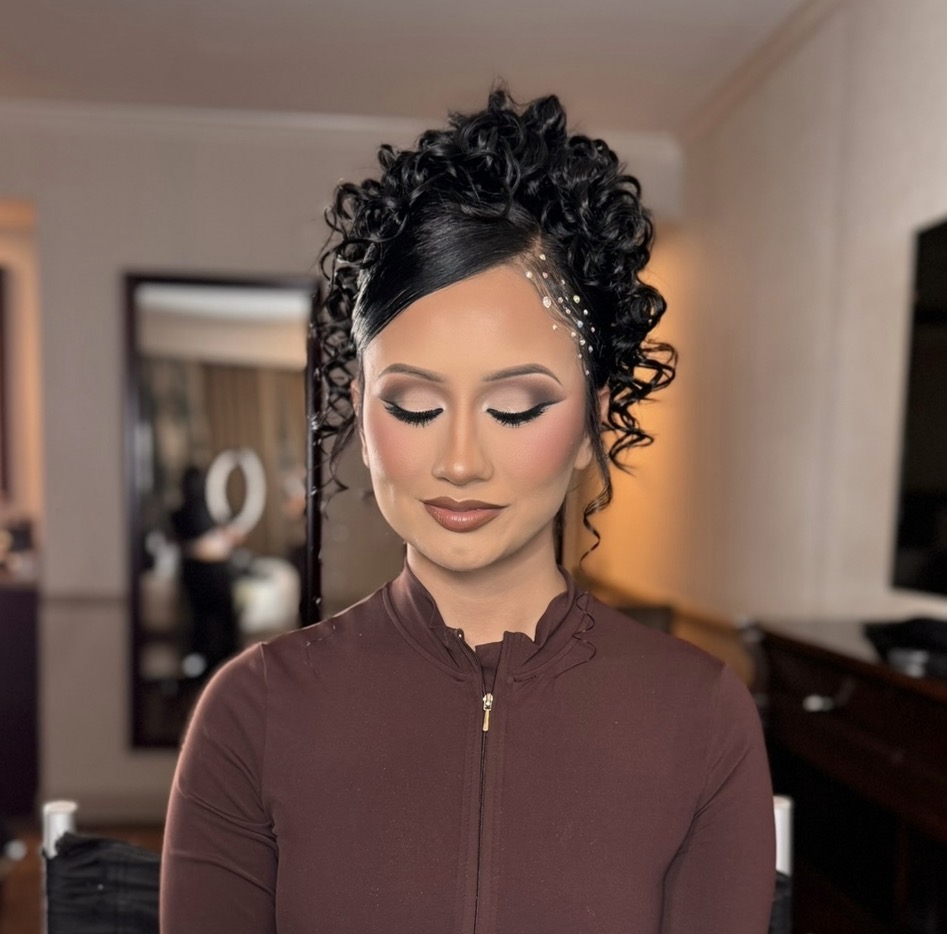 Woman with curly hair and makeup, wearing a brown zip-up top, indoors with a mirror and furniture in background.