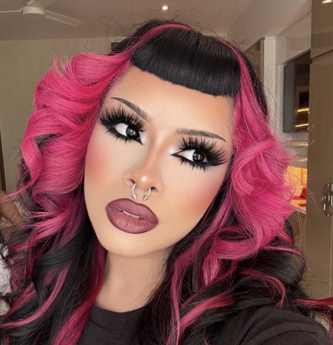 Woman with pink and black wavy hair, bold makeup, and septum piercing, looking at camera indoors.