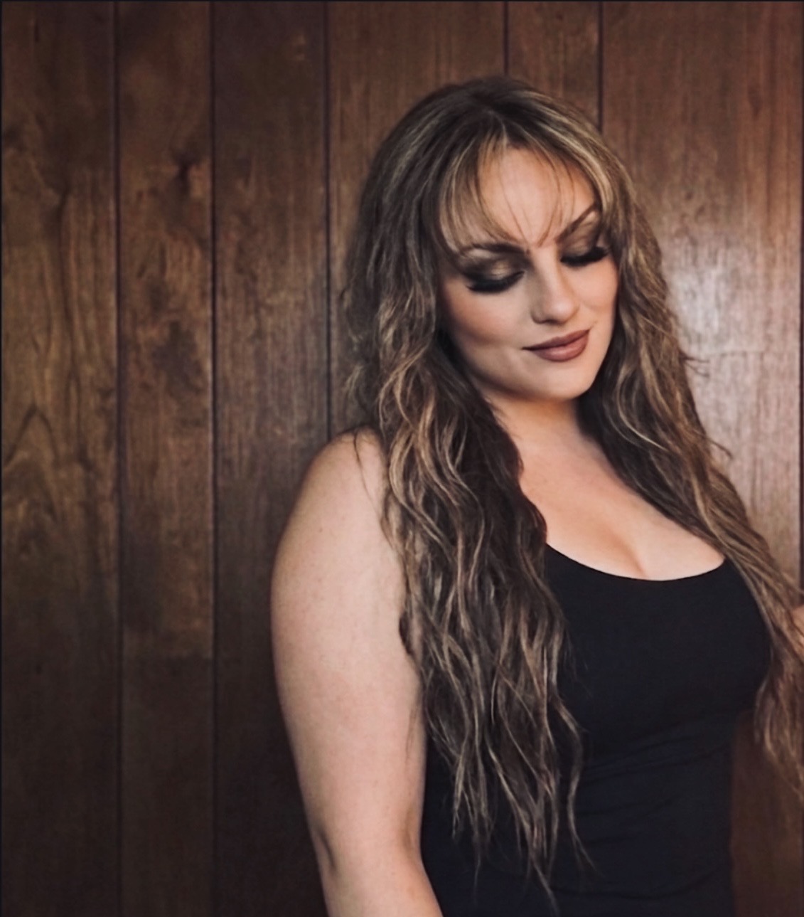 Woman with long wavy hair and makeup, wearing a black top, standing against wooden background.