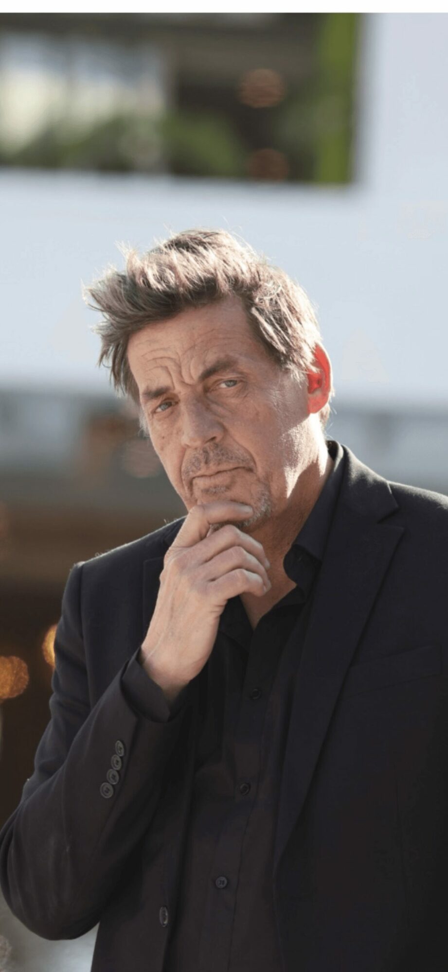 Man with gray hair and beard in black suit, hand on chin, outdoors with blurred background.