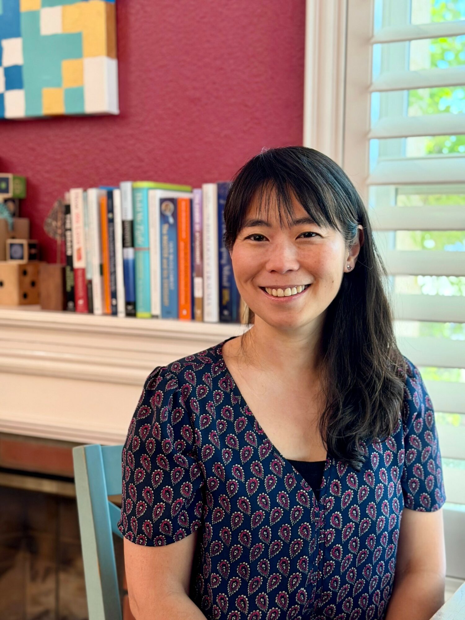 Smiling woman with long dark hair in a patterned dress, sitting indoors near a bookshelf and window with shutters.