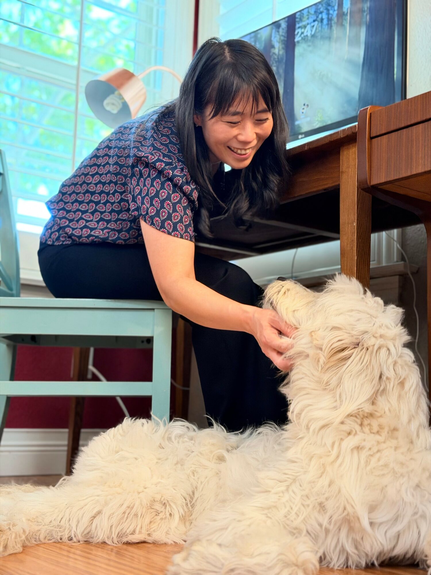 Woman smiling and petting a large, fluffy dog indoors near a desk and window.
