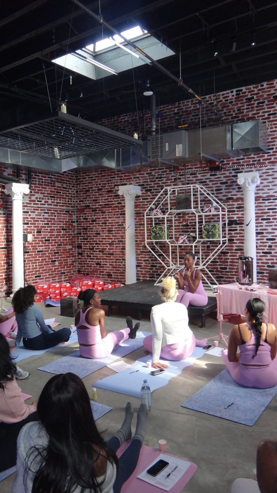 Group of people sitting at tables in a brick-walled indoor space with large windows and a decorative metal structure.