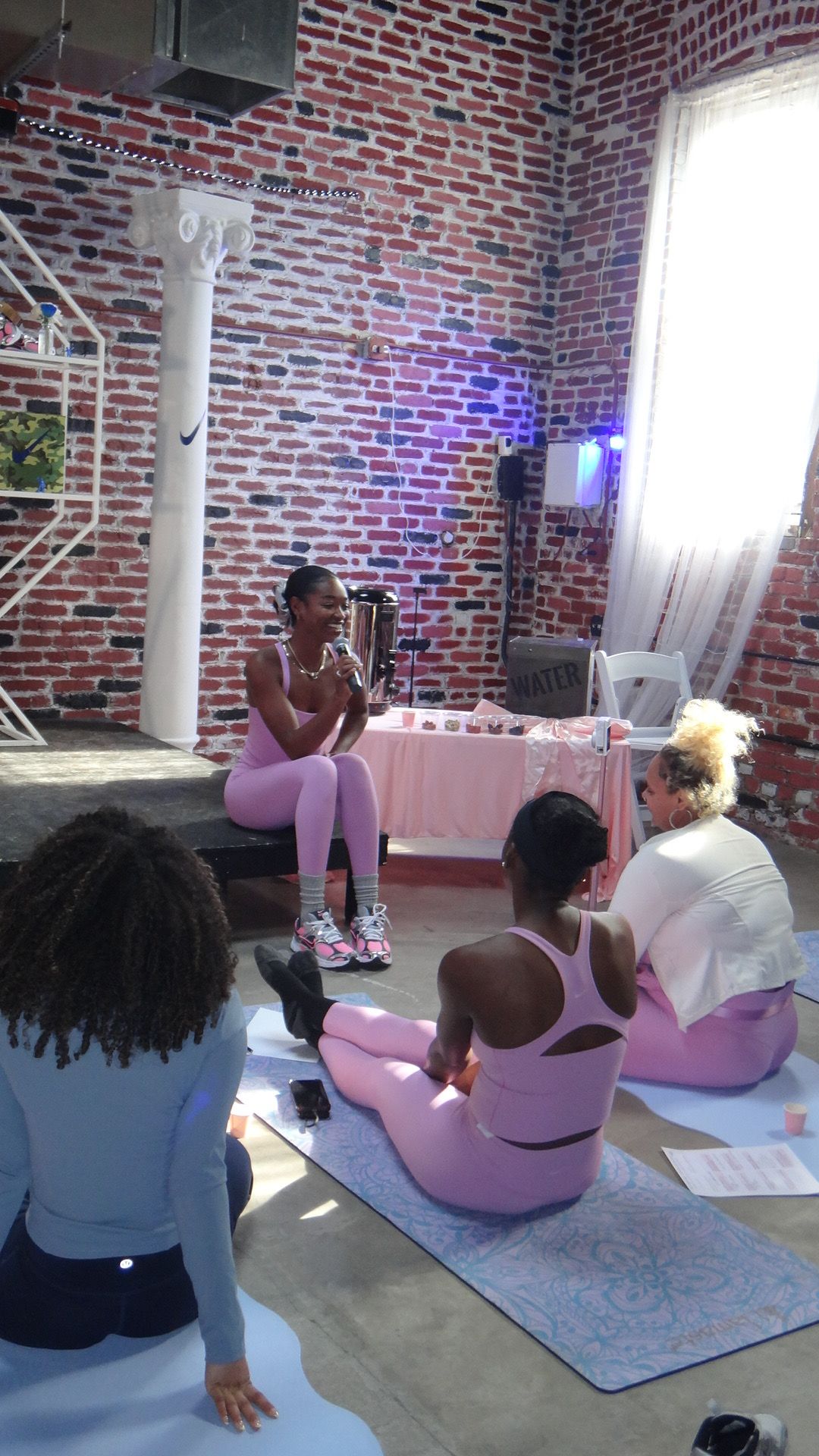 People sitting on pink chairs in a room with brick walls and a white column, some standing and talking.