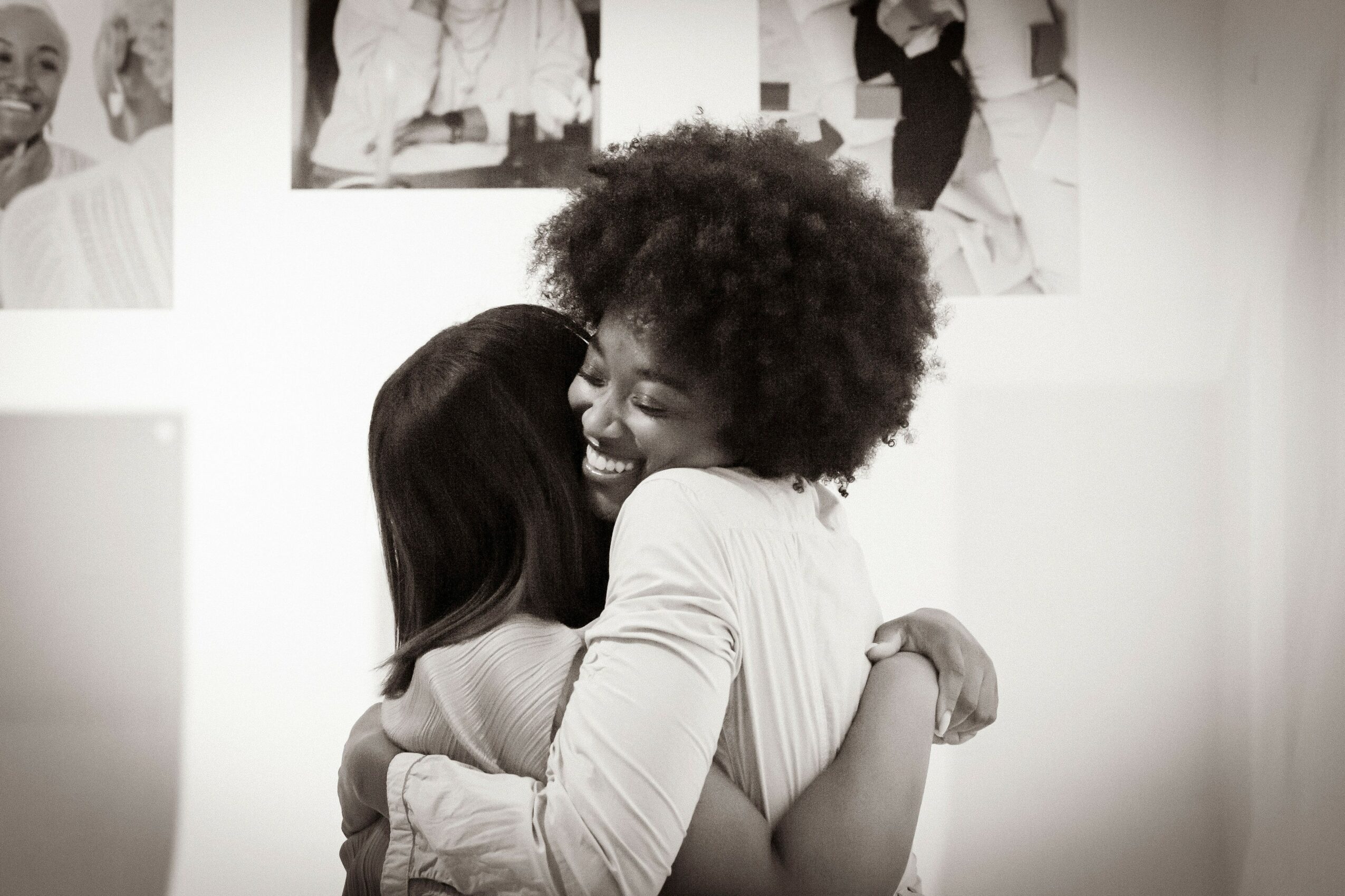 Two women hugging and smiling, one with curly hair and the other with straight hair, in a room with photos on the wall.