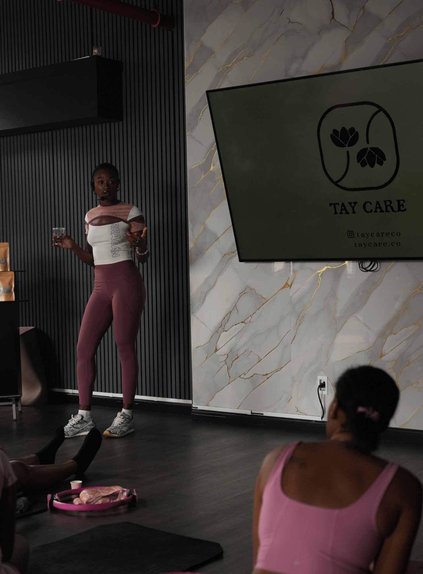 Person standing and speaking in front of a group, with a large screen displaying a logo and text, in a modern room.