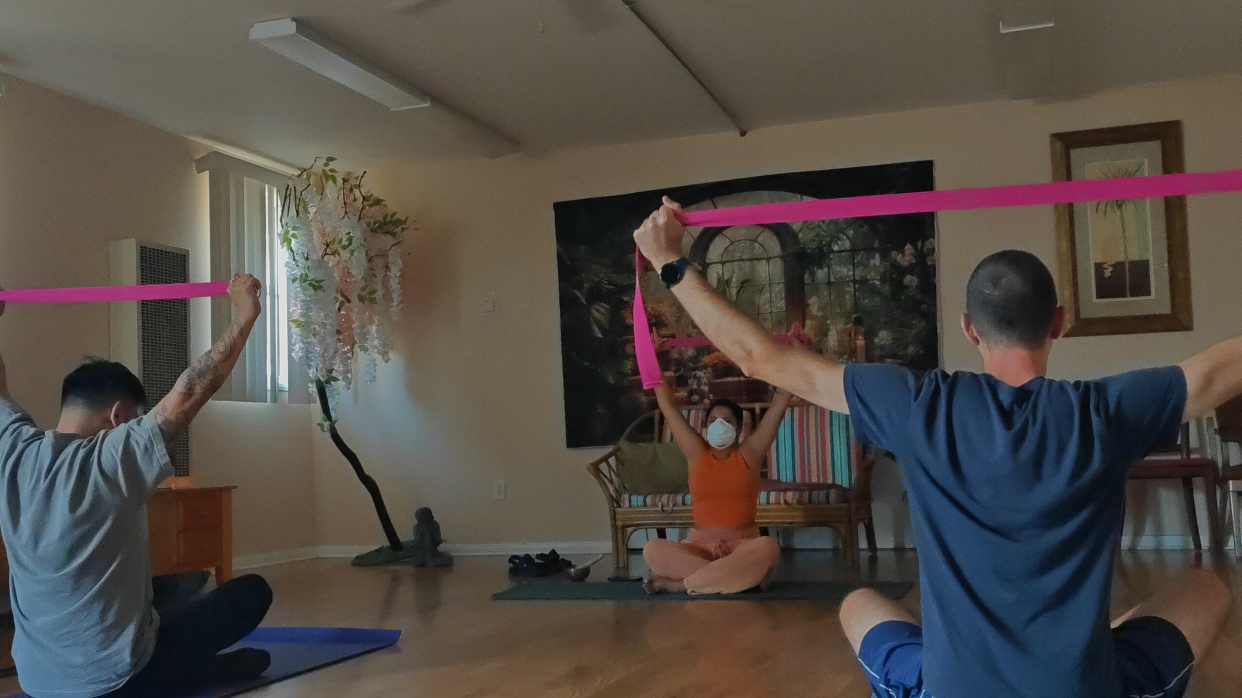 Three people practicing yoga with pink resistance bands in a room with a large window and artwork.