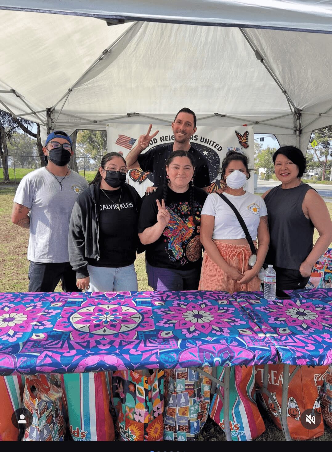 Group of seven people standing under a tent, smiling, with a colorful table in front, outdoors on a sunny day.